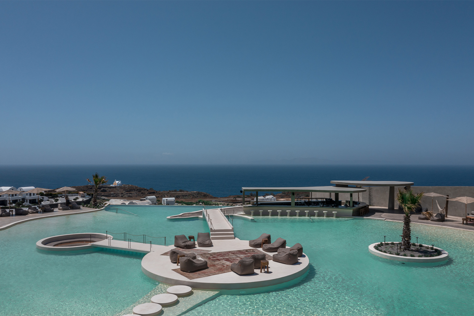 A large pool area surrounded by sun loungers and a bar, in the cente of the pool is a stone platform where there are more sunloungers, the ocean is seen in the background
