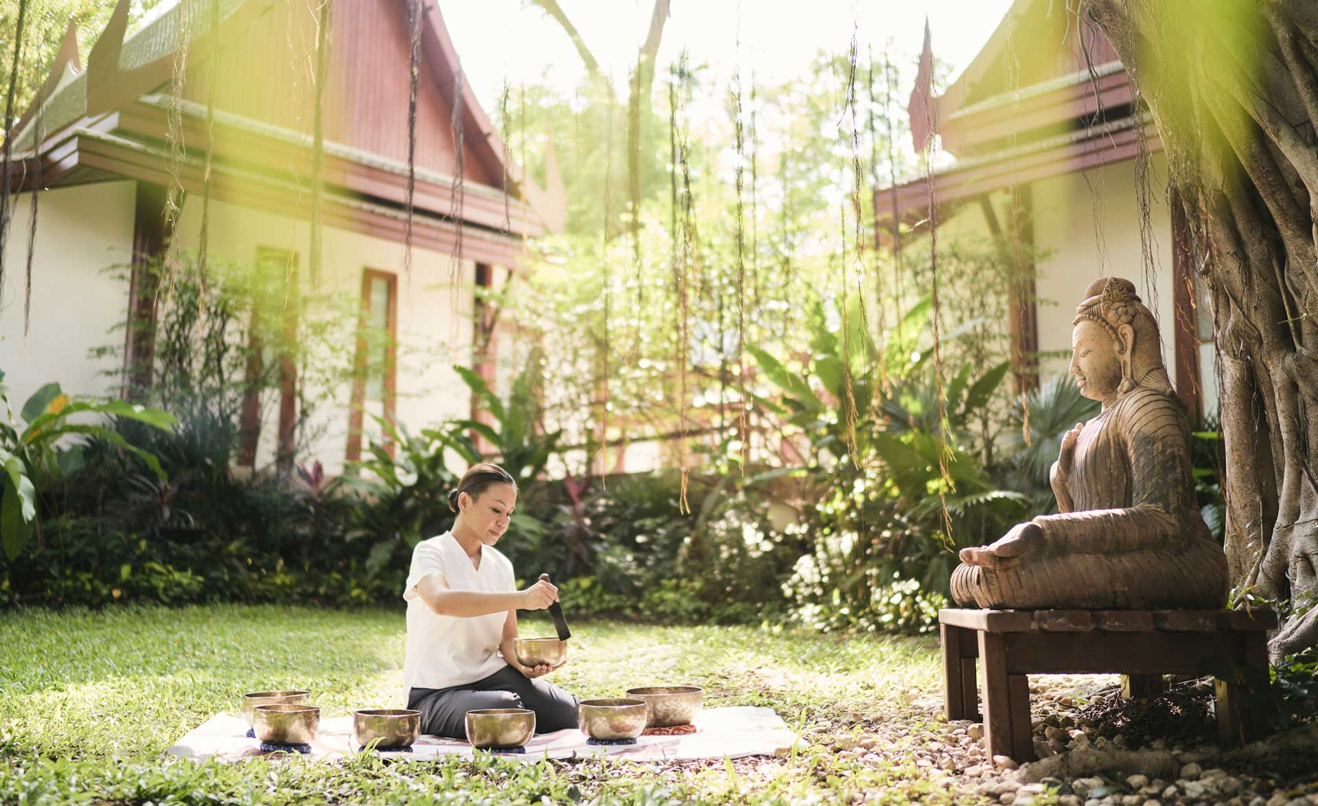 Woman sitting on a mat outdoors, playing Tibetan singing bowls near a statue in a lush garden.