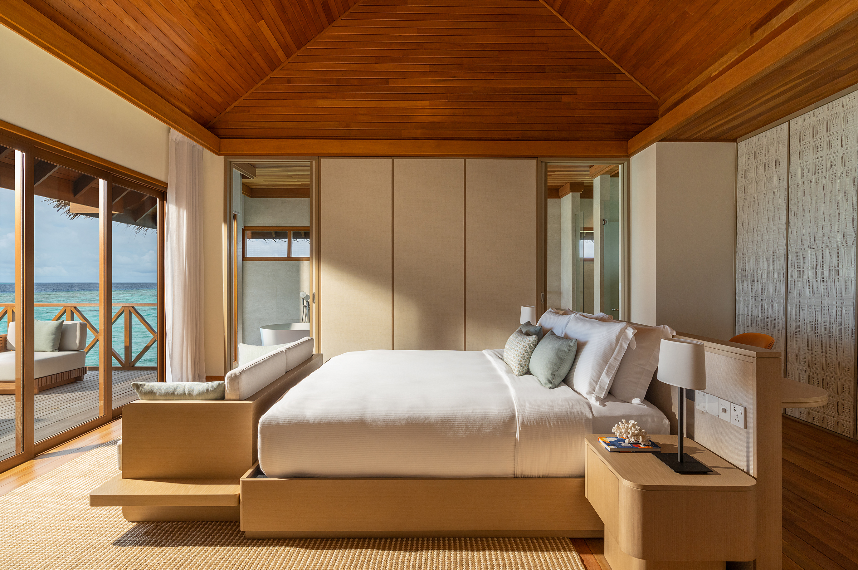 The master bedroom of an Ocean Pavilion at Huvafen Fushi with neutral decor under a beamed ceiling and a bed facing large sliding doors onto a deck