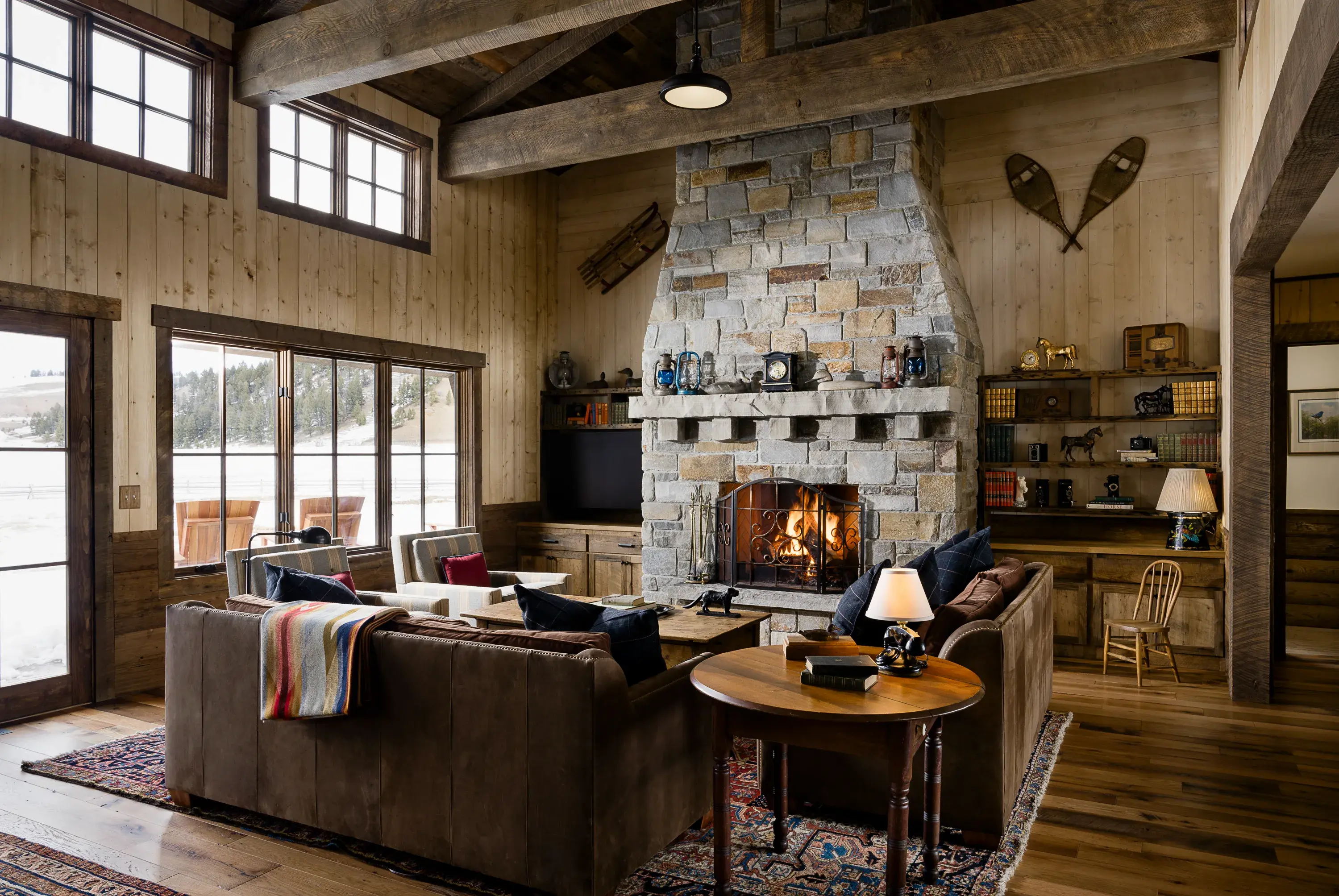 Living room of Red Tail Cabin at The Ranch at Rock Creek in Montana featuring a large stone fireplace with cosy ranch-style furniture and decorative shelving with large windows looking out onto a snowy landscape