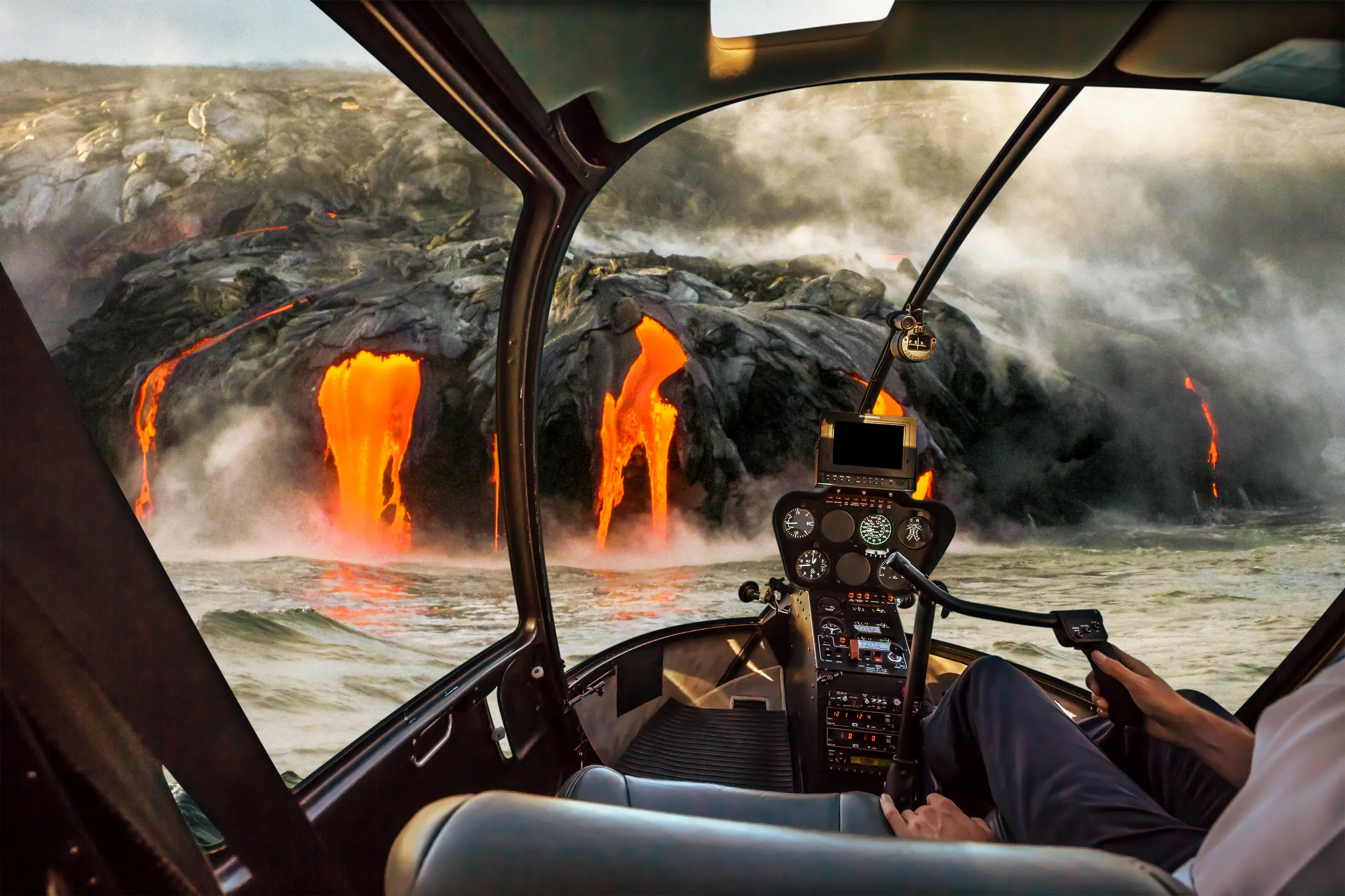 The side of an erupting volcano flowing into the ocean from the inside of a helicopter with a glimpse of controls and a pilot