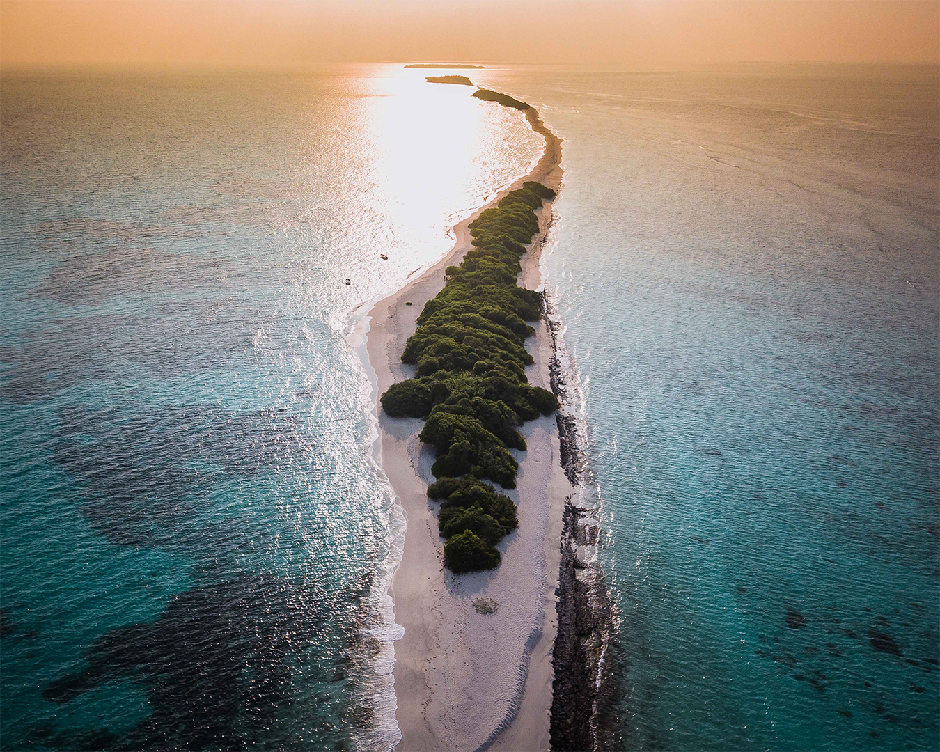 Aerial view of a narrow strip of land with lush greenery surrounded by calm blue waters at sunset.