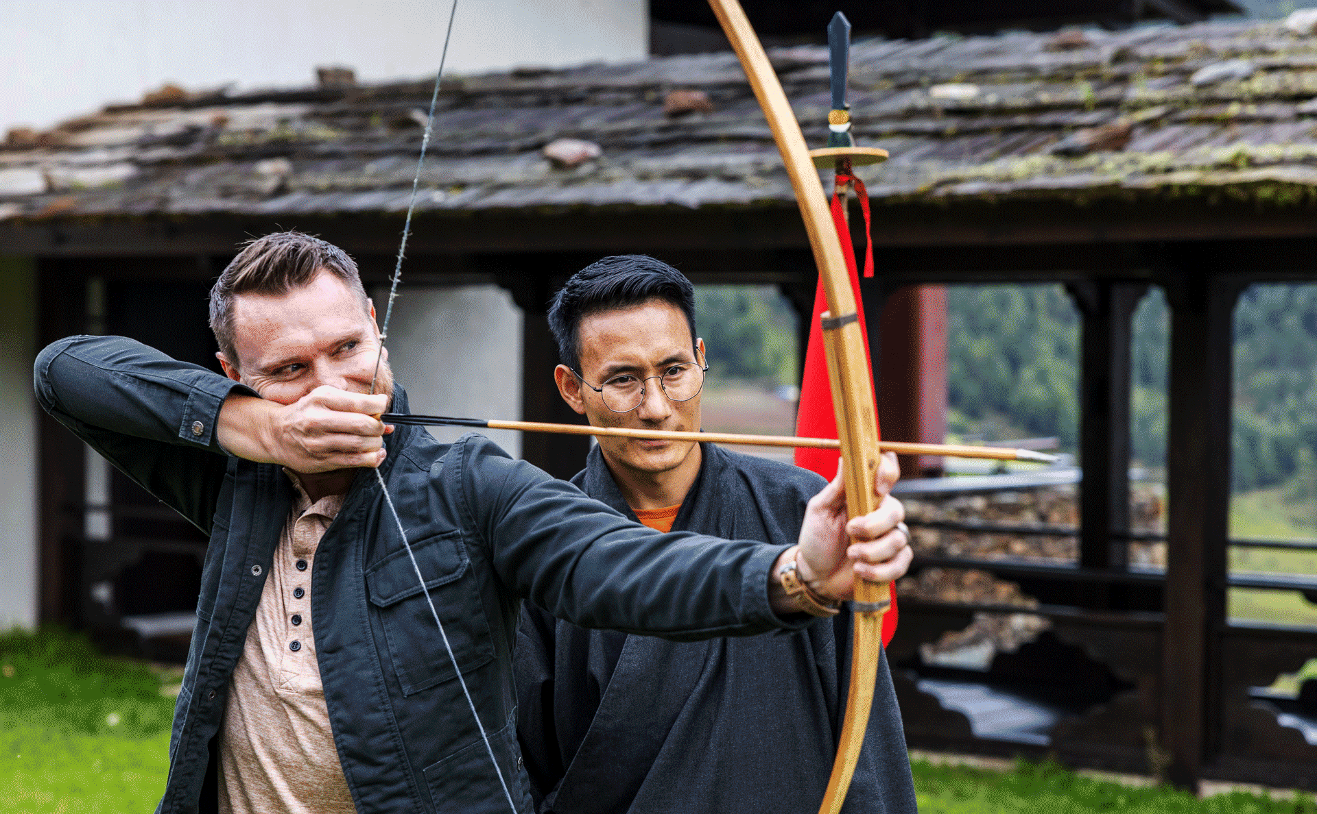 Two people practising archery outdoors, with one guiding the other’s stance beside a traditional building.