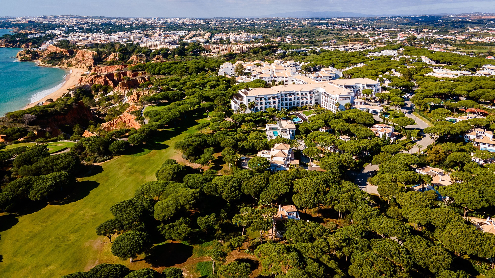 An aerial view of a coastal resort at Pine Cliffs Portugal, with a beach, cliffs, and numerous buildings surrounded by dense green trees