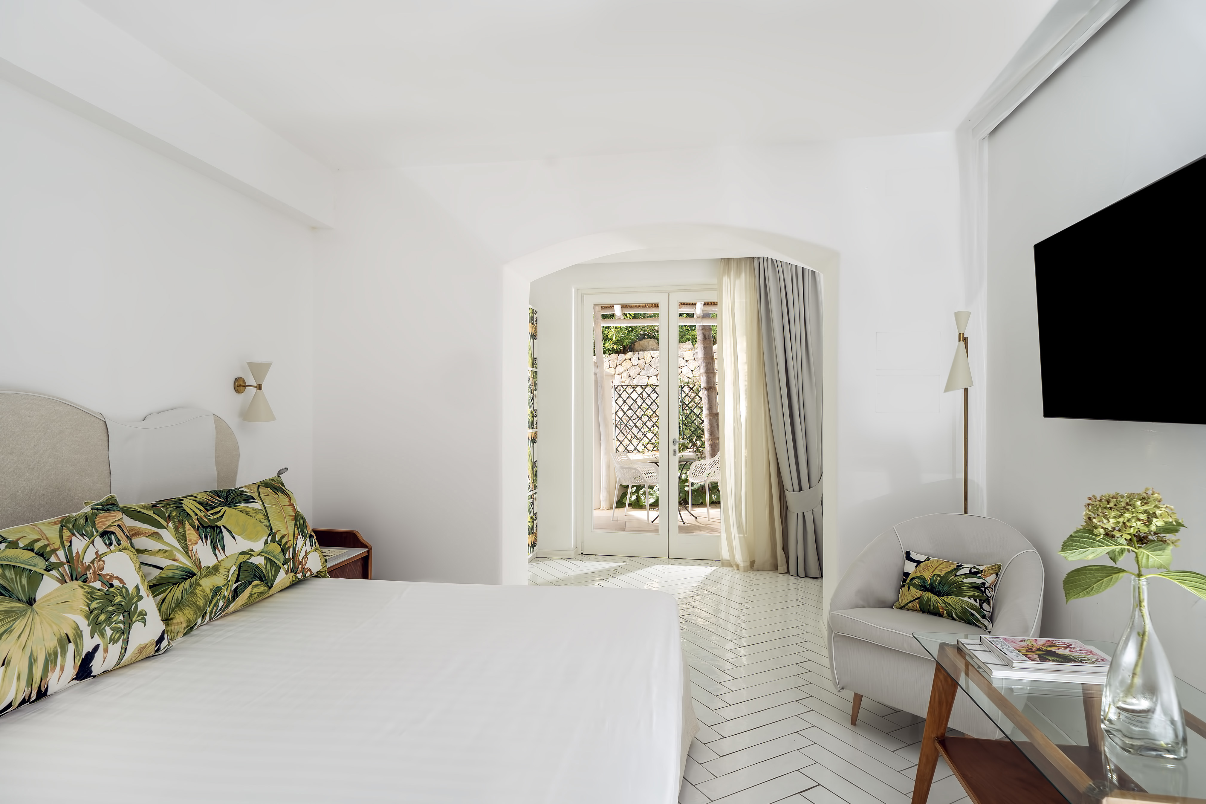 Bright and airy hotel room at Botania Relais & Spa in Ischia featuring a comfortable bed with tropical-themed pillows, a cosy seating area, and a view of the garden through French doors.
