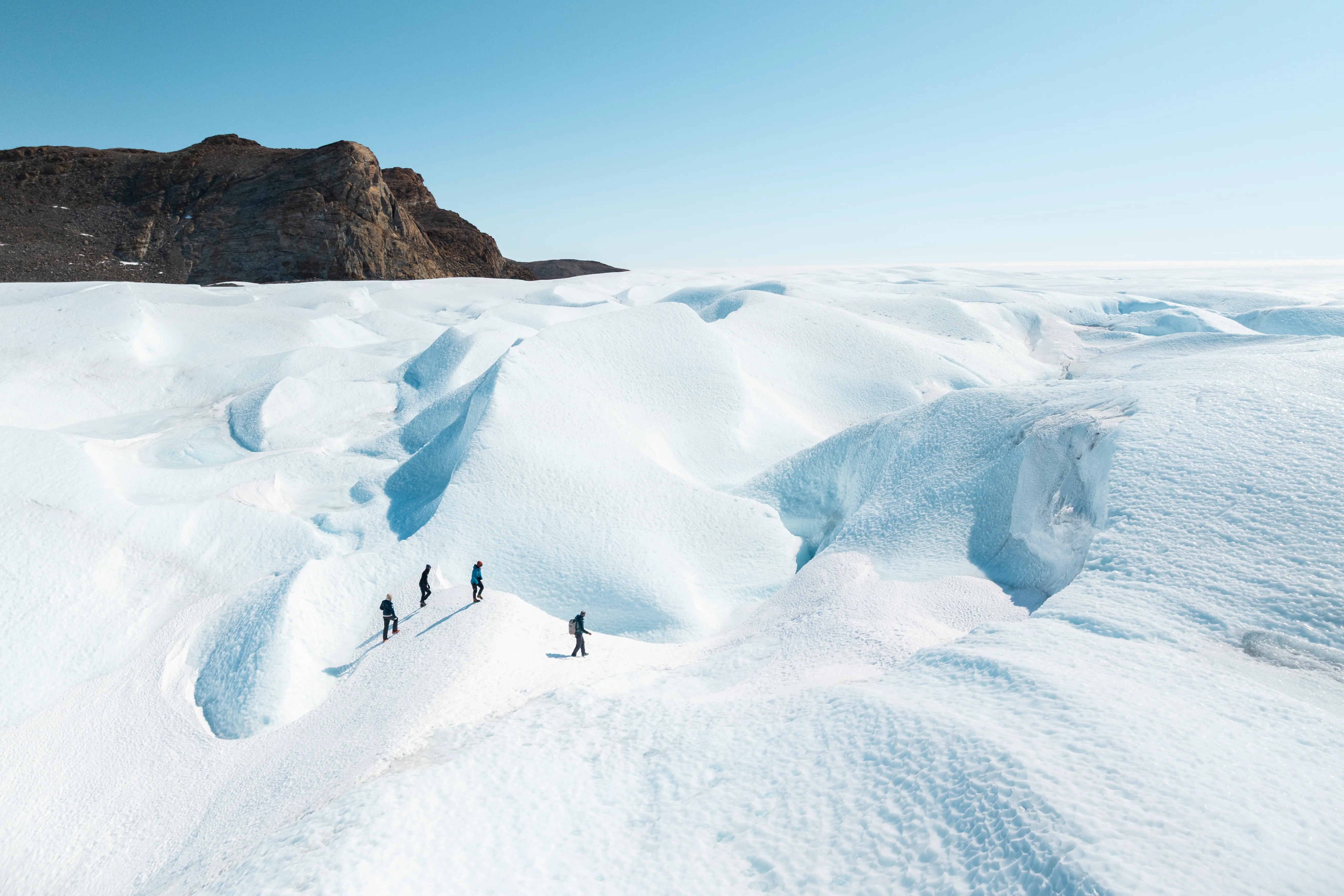 Four people trekking across a bright, snow‑covered Antarctic landscape with rugged ice formations under a clear blue sky.