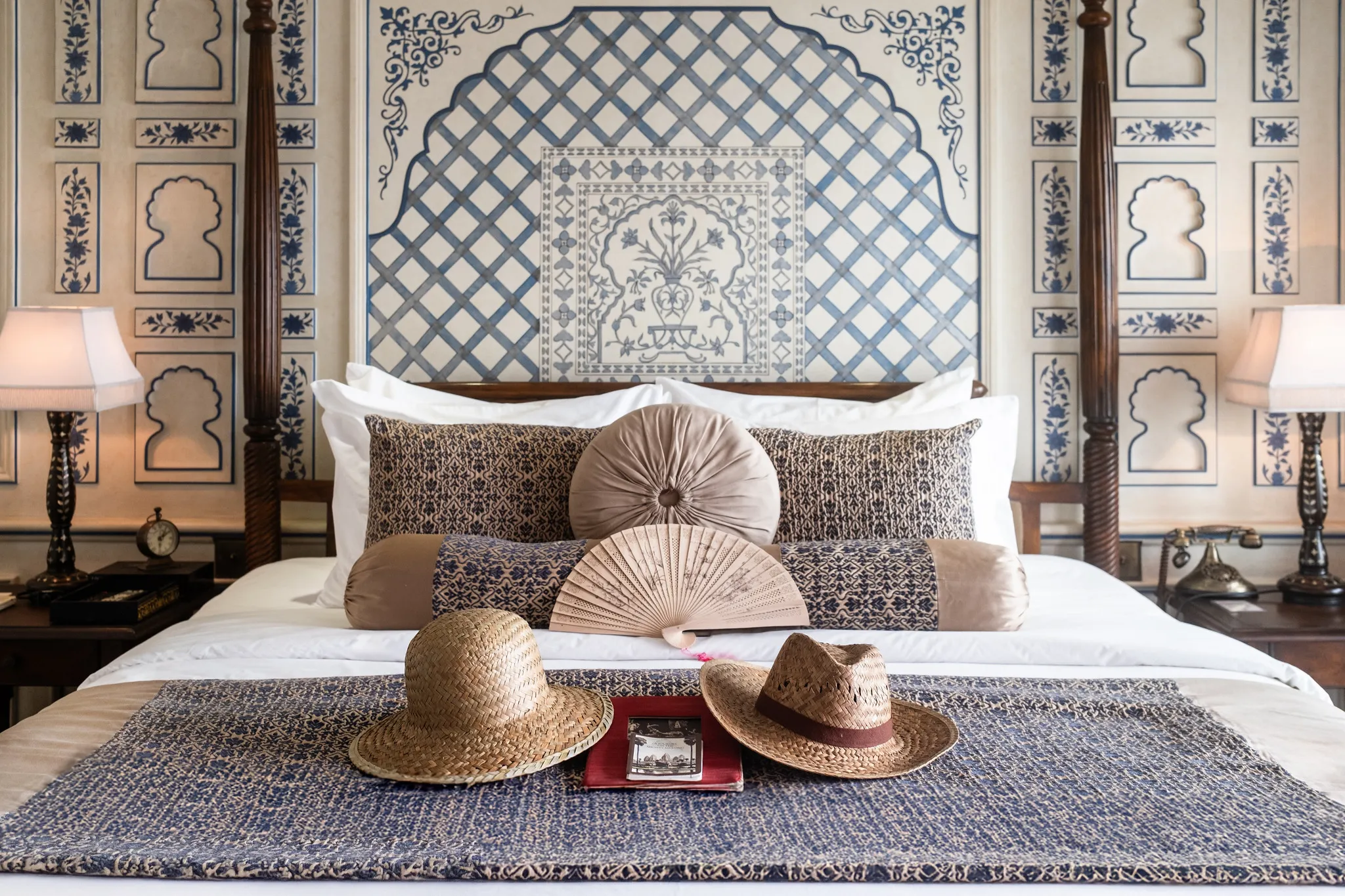 A luxurious Heritage Line cruise cabin featuring a bed with decorative pillows, a fan, and two hats on a woven blanket, set against a blue and white patterned wall.