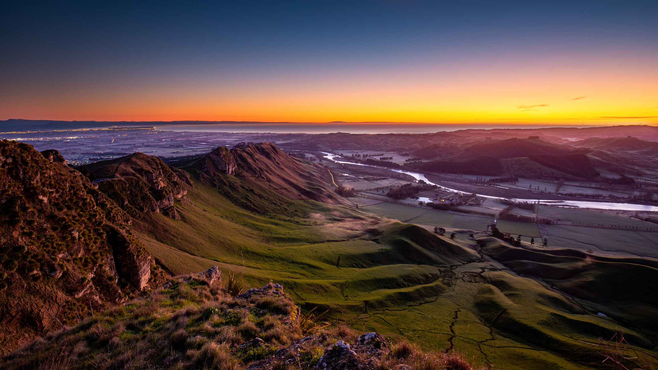 A scenic view of a green valley with rolling hills and a river at sunset, with the sky transitioning from orange near the horizon to deep blue higher up