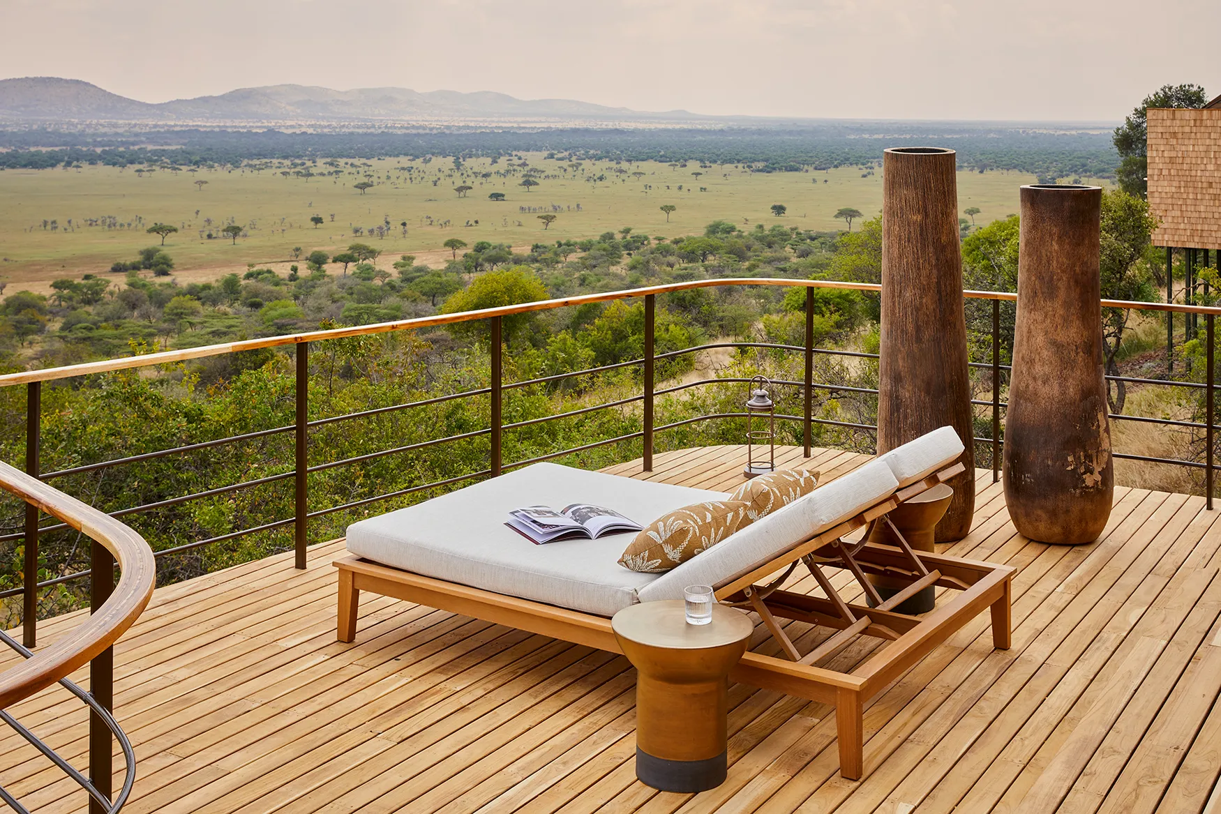 Elevated wooden deck with a cushioned daybed overlooking a sweeping savannah landscape and distant hills.