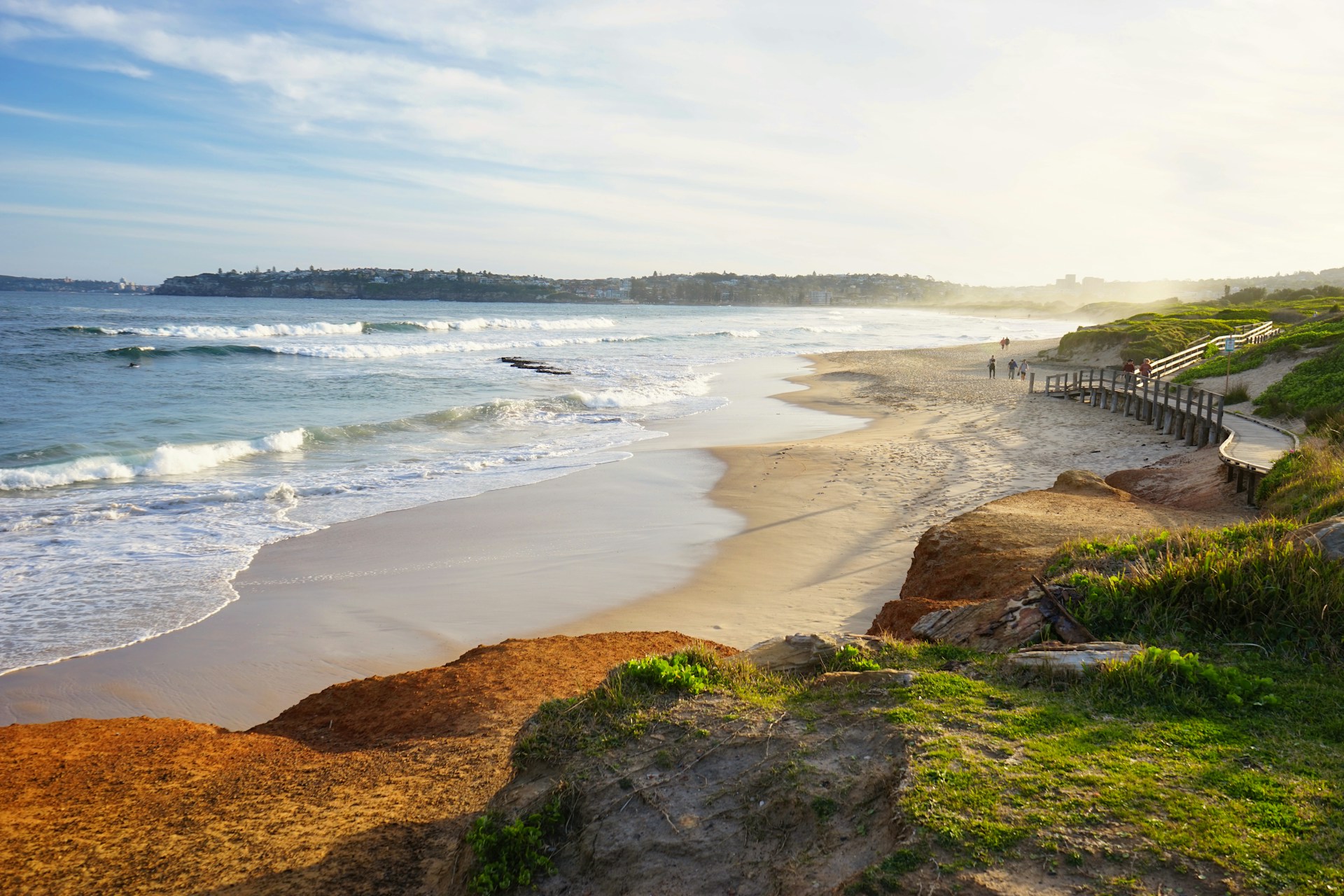 A scenic coastal landscape with rocky cliffs, lush green hills, and waves crashing against the shore under a partly cloudy blue sky