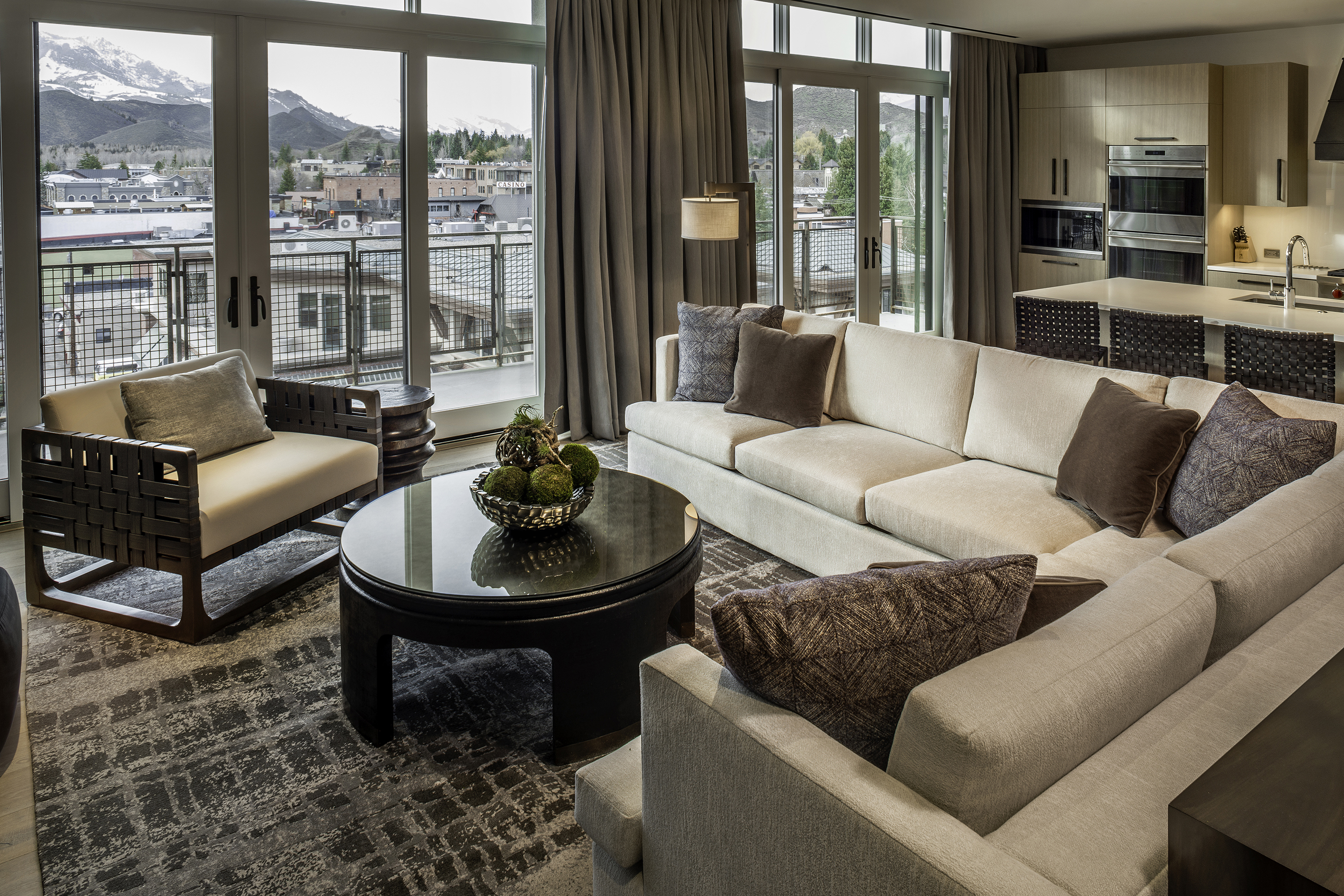 A modern living room at Limelight Ketchum hotel with a beige sectional sofa, round coffee table, armchair, and floor-to-ceiling windows showcasing mountain and town views