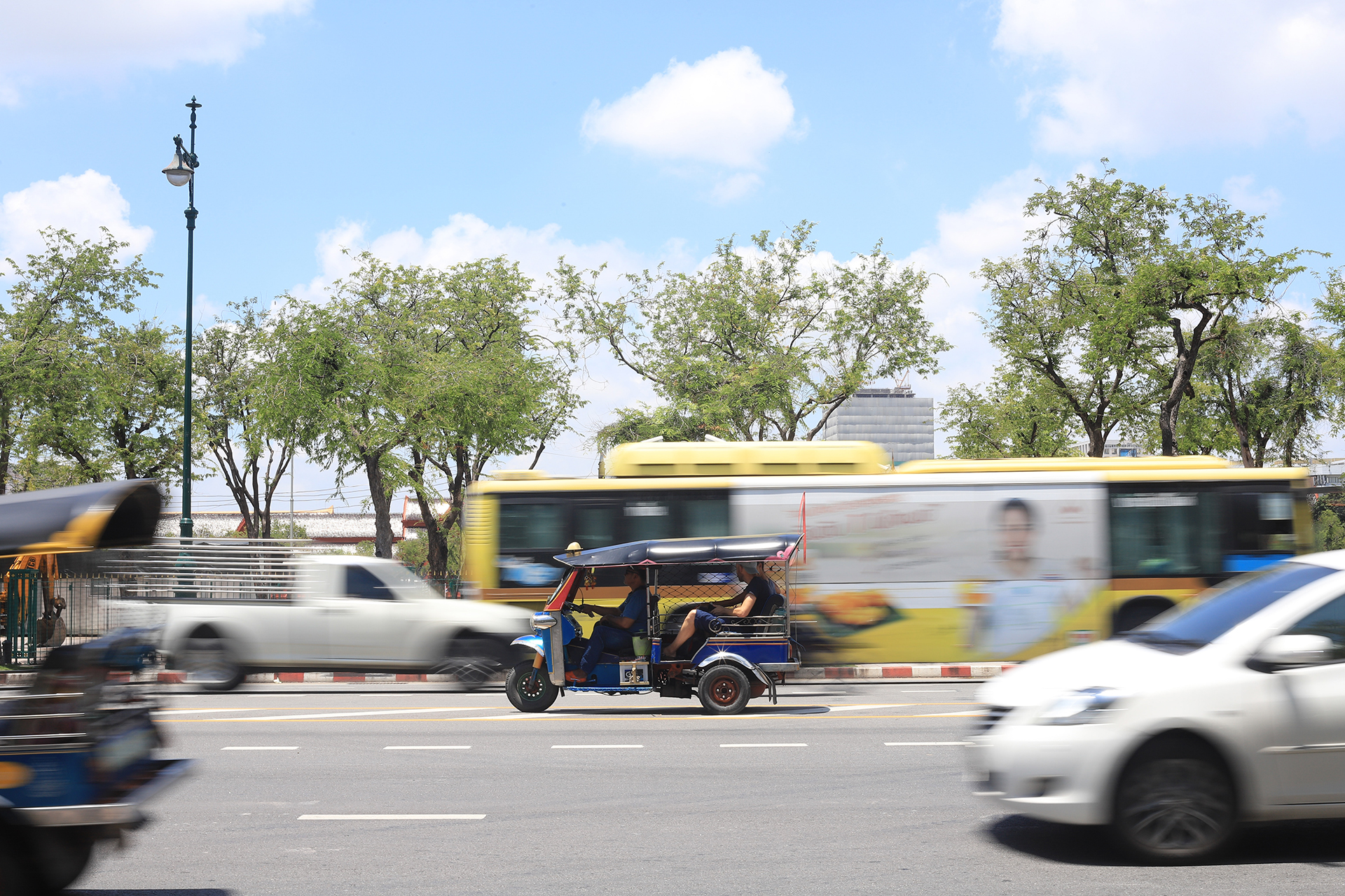 A busy road with a bus and cars out of focus but an in focus tuk tuk is center of the image