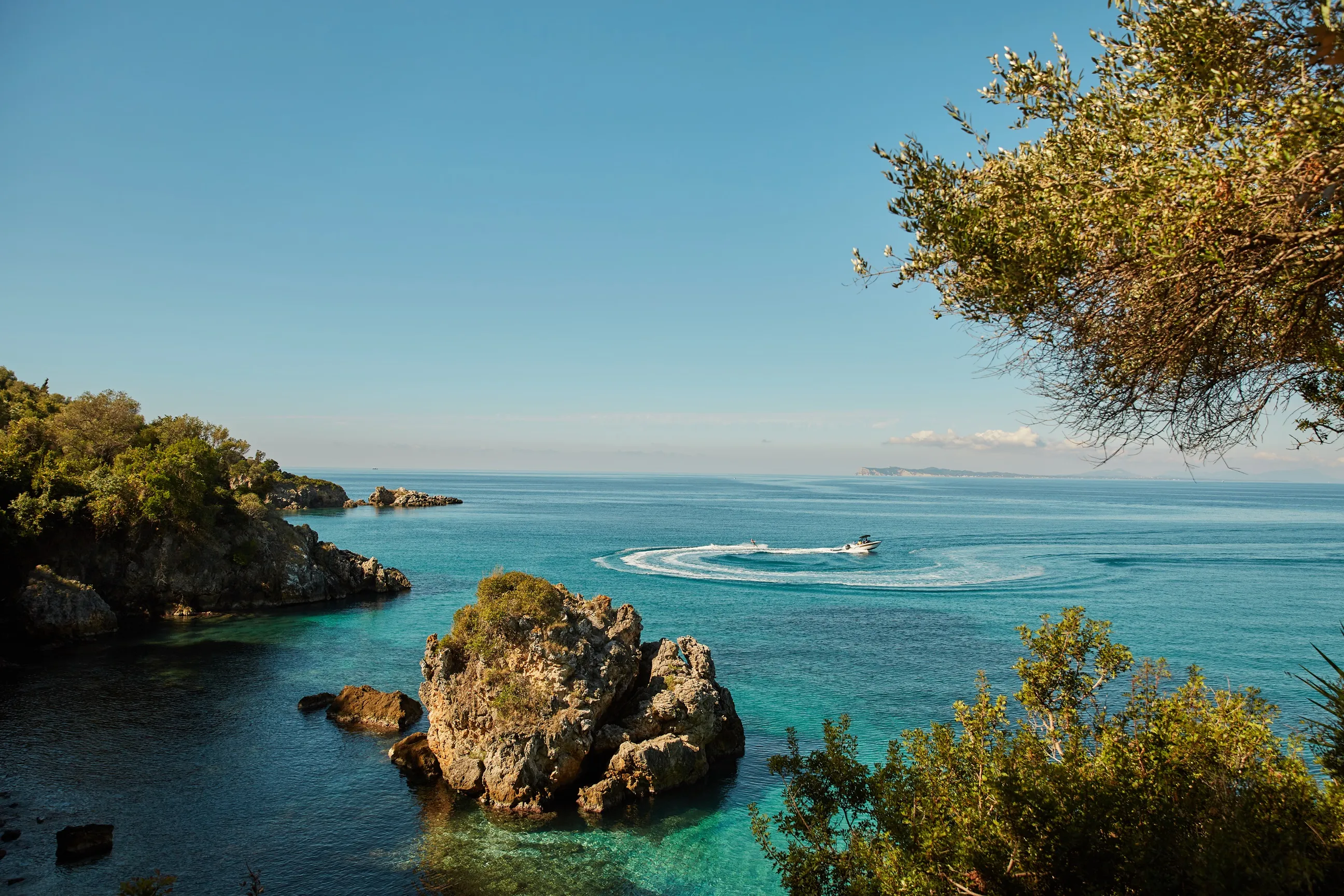 Scenic coastline with clear blue water, rocky outcrops, and a boat making a circular wake