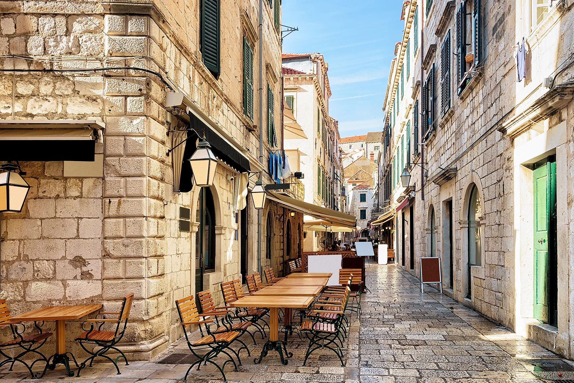 A stone street in Dubrovnik featuring a closed restaurants with light wooden tables and chairs set out on the pavement