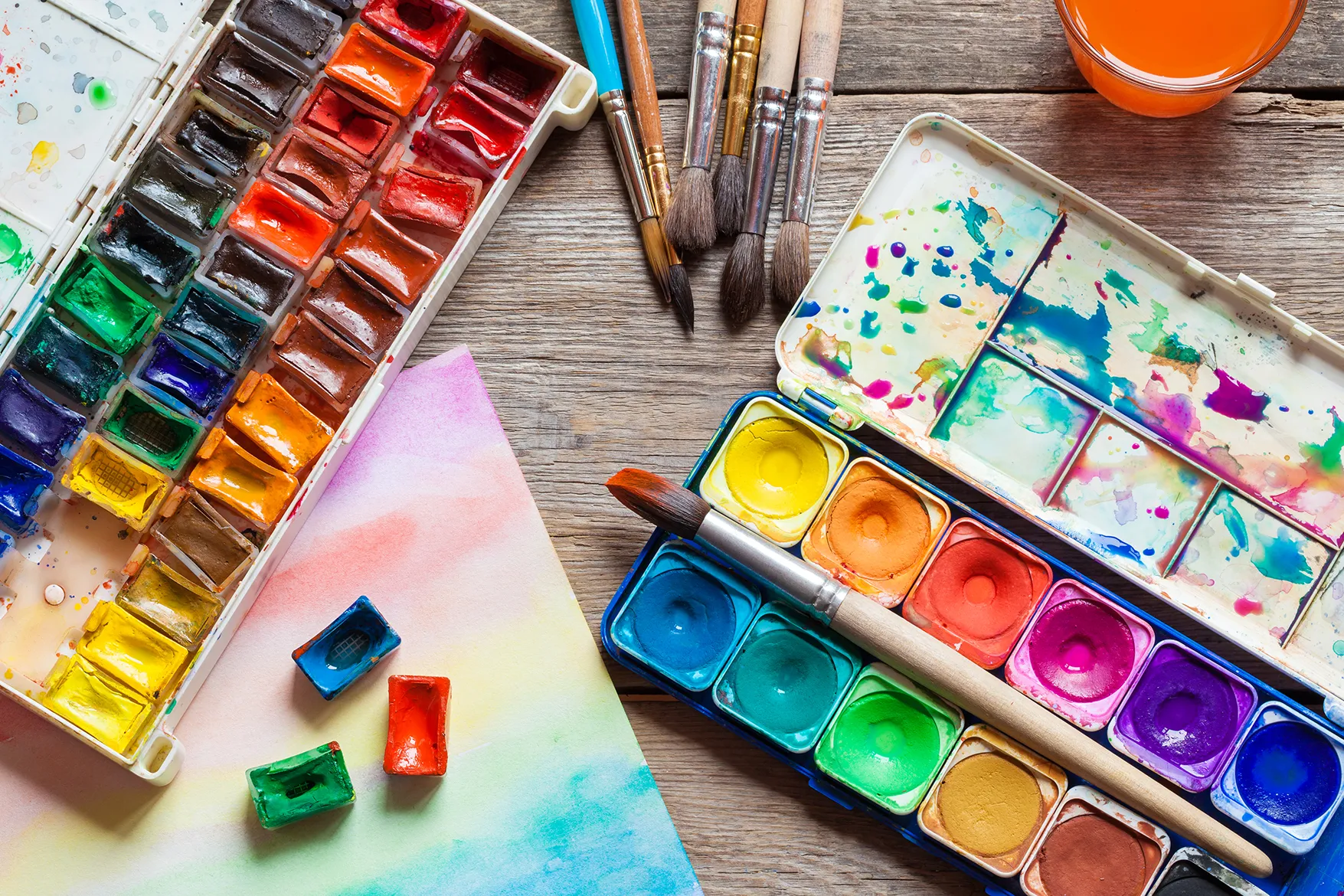 Watercolor paint sets, brushes, and paper arranged on a wooden table with vibrant splashes of color.