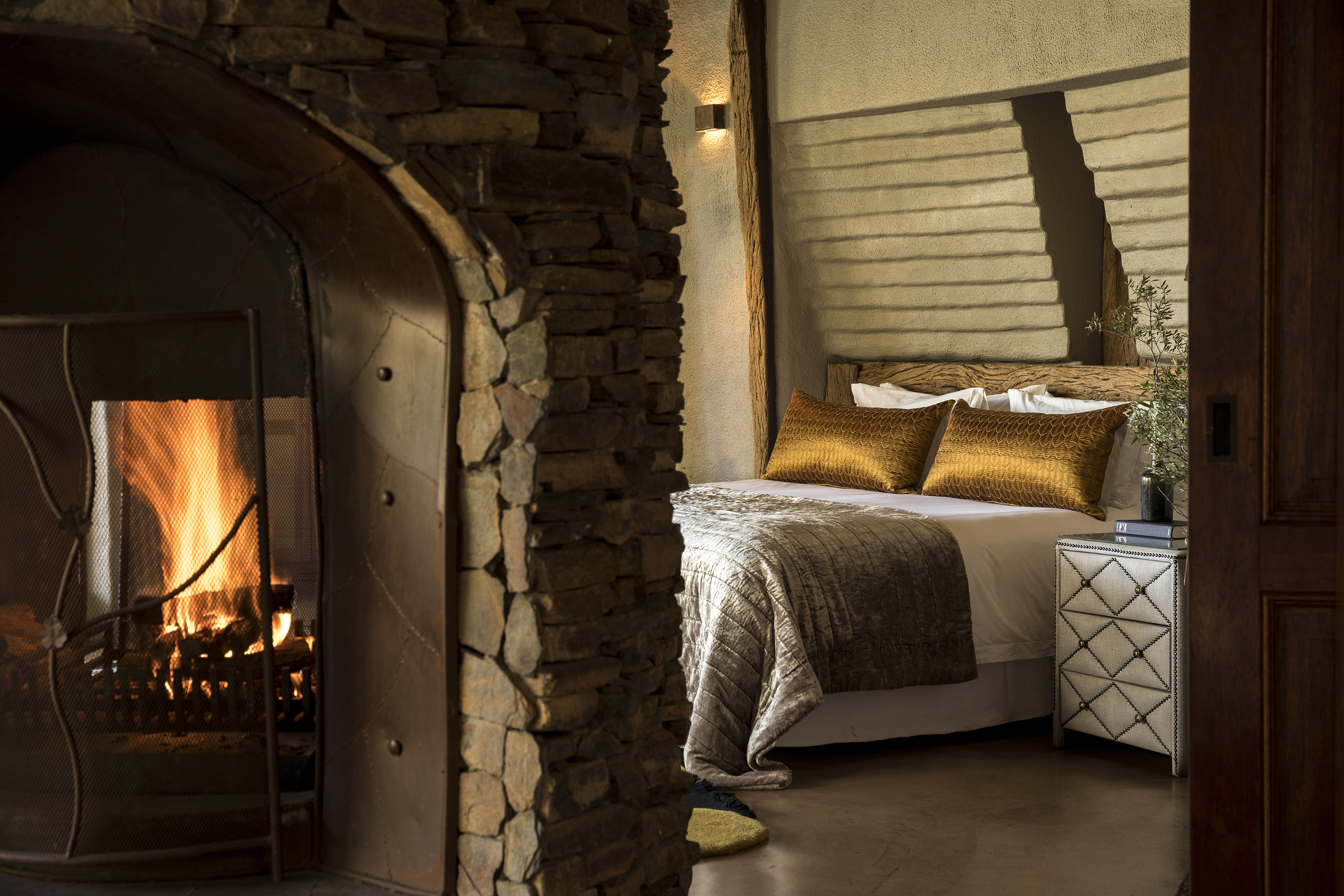 A lit stone fireplace with the bed of a suite further behind that features bronze pillows and a comfy throw