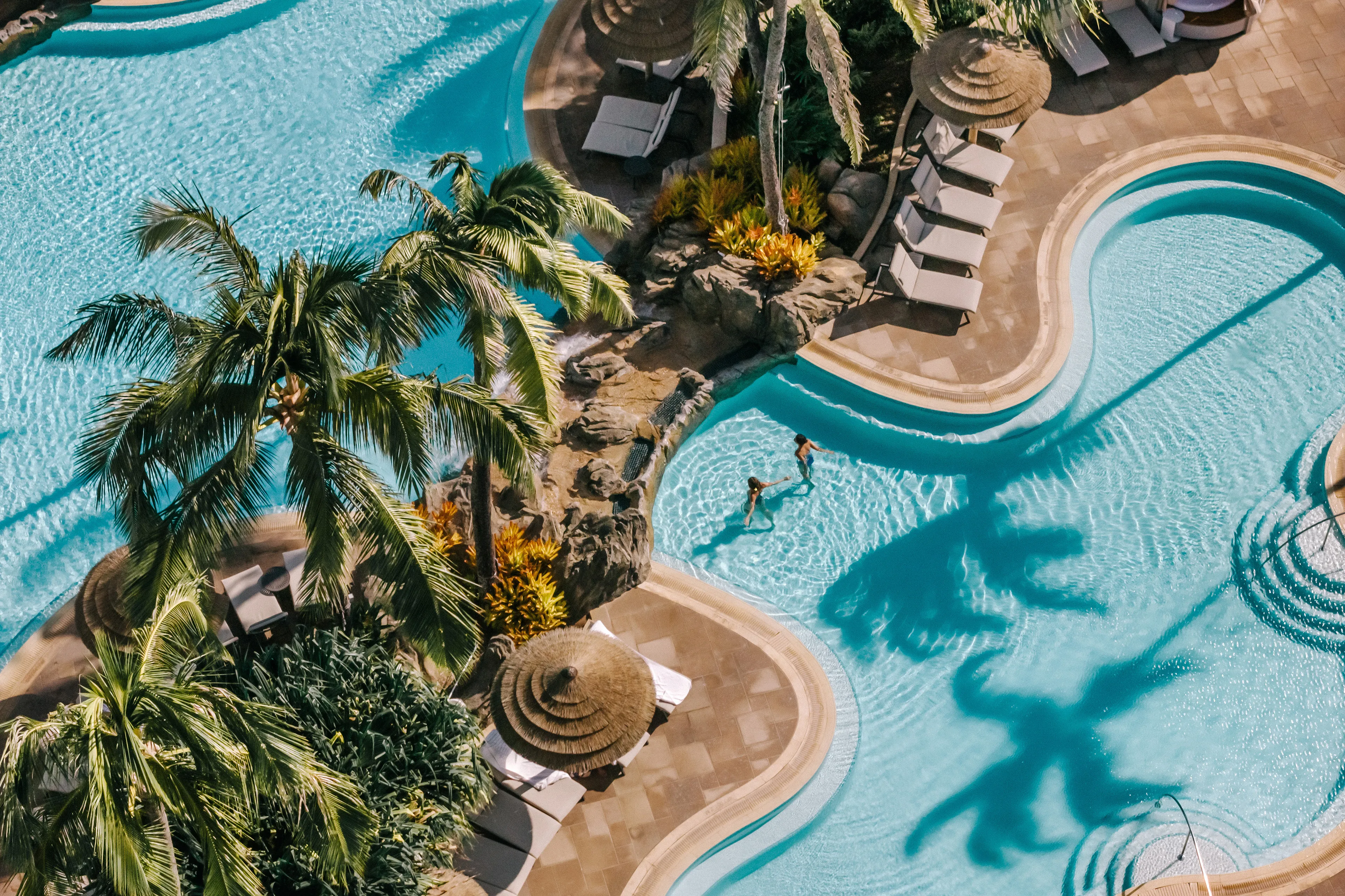 Two guests in the pool at The Ritz-Carlton Maui, Kapalua from above, featuring palm trees and thatched sun umbrellas on the side