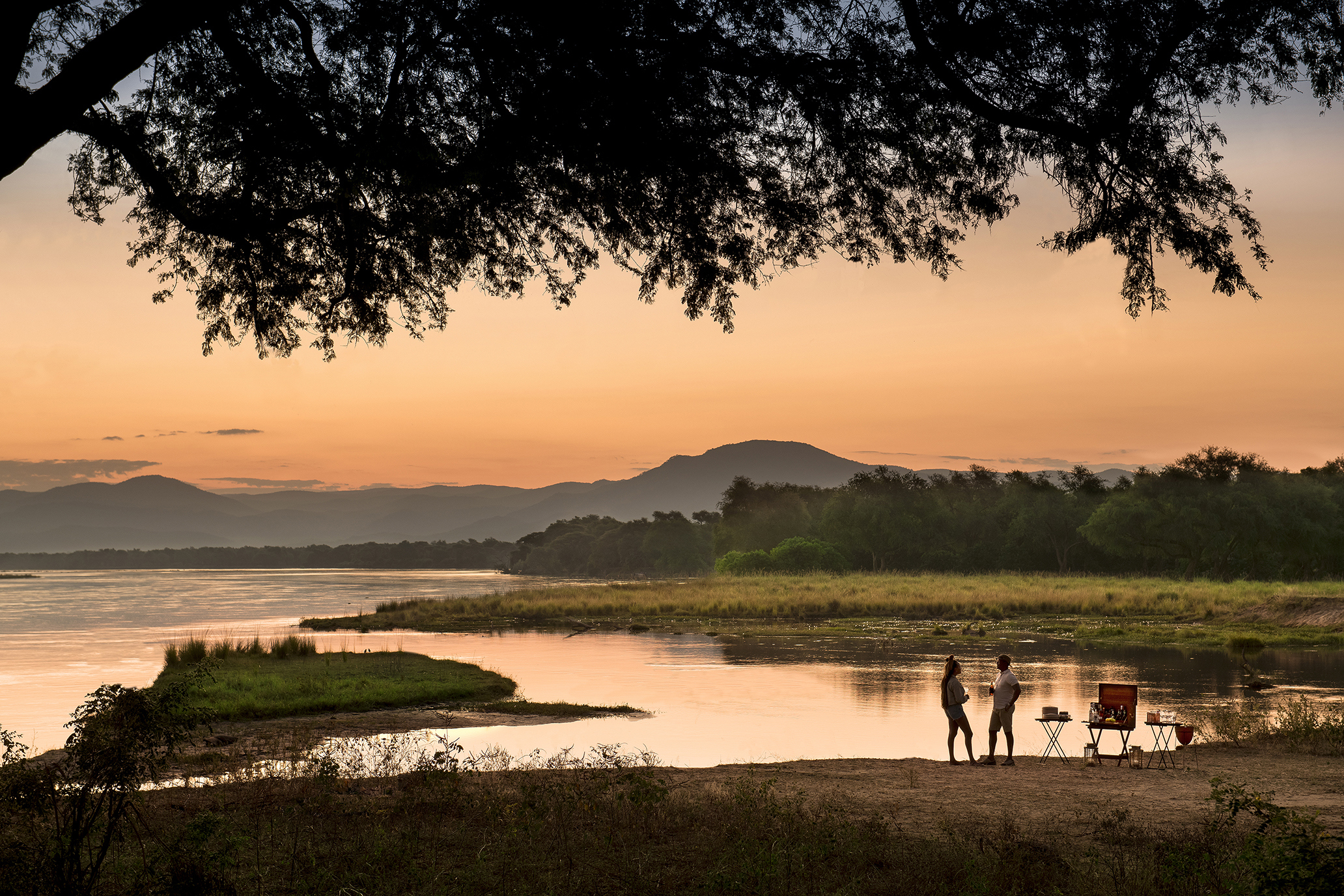 A couple enjoying drinks during sunset by the Zambezi River under an acacia tree