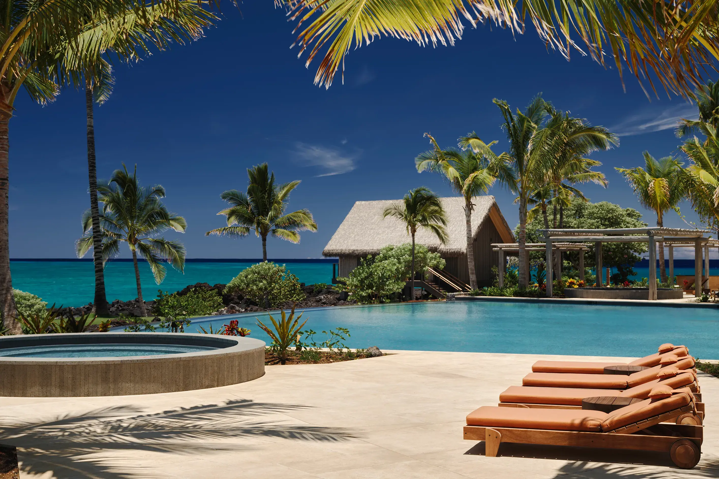 A pool and hot tub at Rosewood Kona Village with orange sun loungers and a thatched building framed by palms