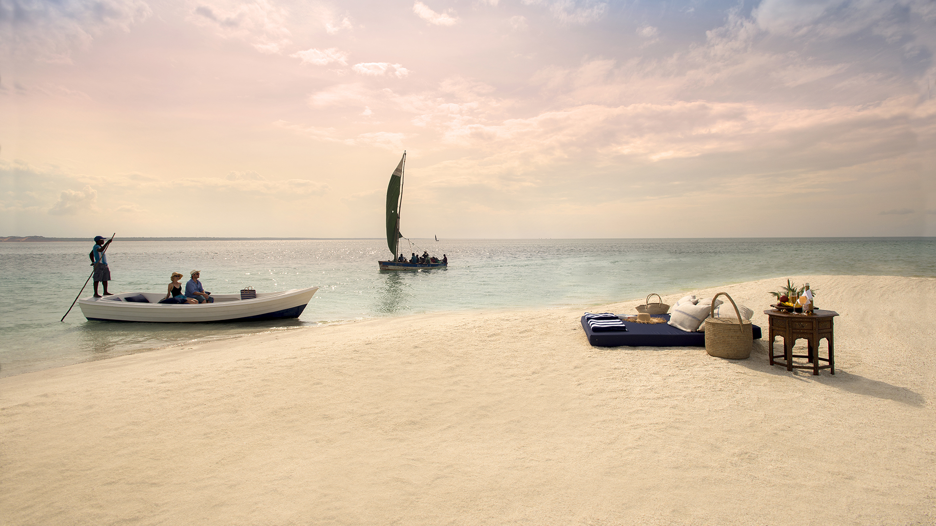 Two small boats approach the shore of a white sand beach where a lavish picnic area awaits them