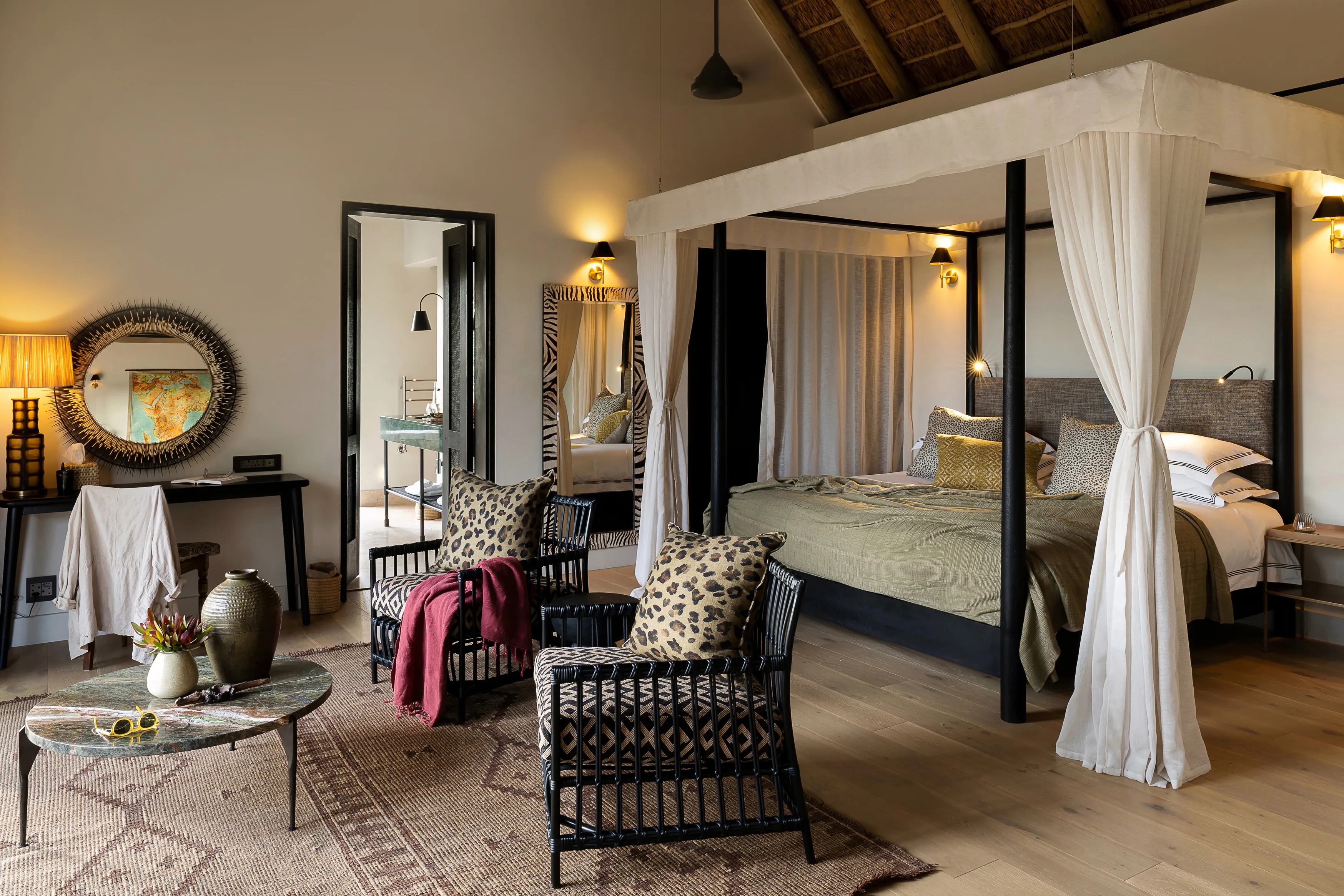 The cosy interior of a suite at Monwana Game Reserve featuring two chairs with leopard print cushions, a marble coffee table with vases, a vanity area and large four poster bed with mesh curtains and khaki blankets