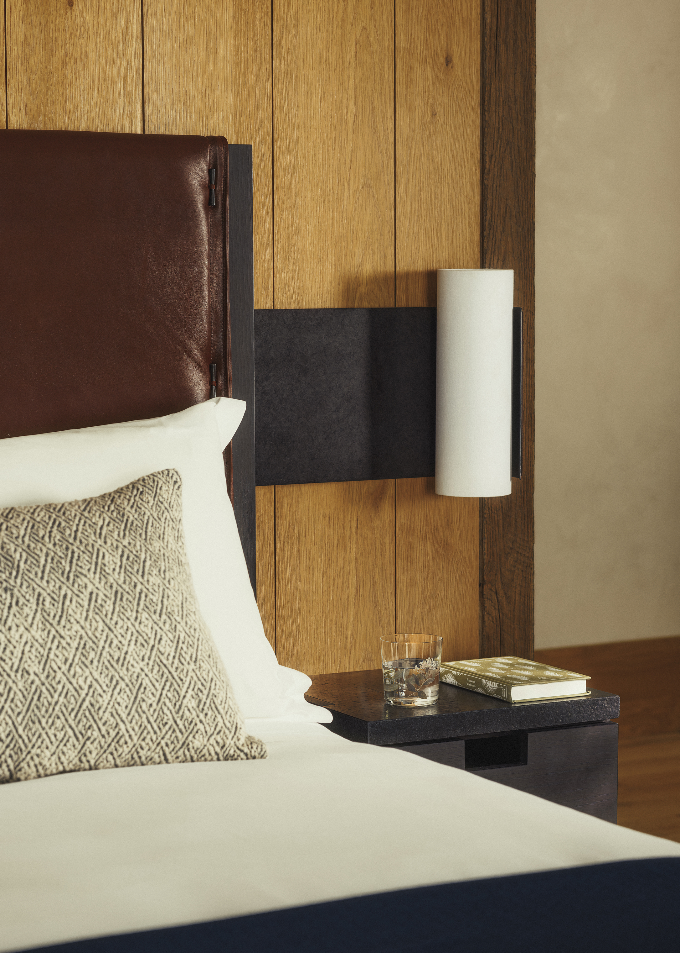 A close up on a bedside table featuring a book and glass below a tubular light with a glimpse of a comfy bed with cream sheets