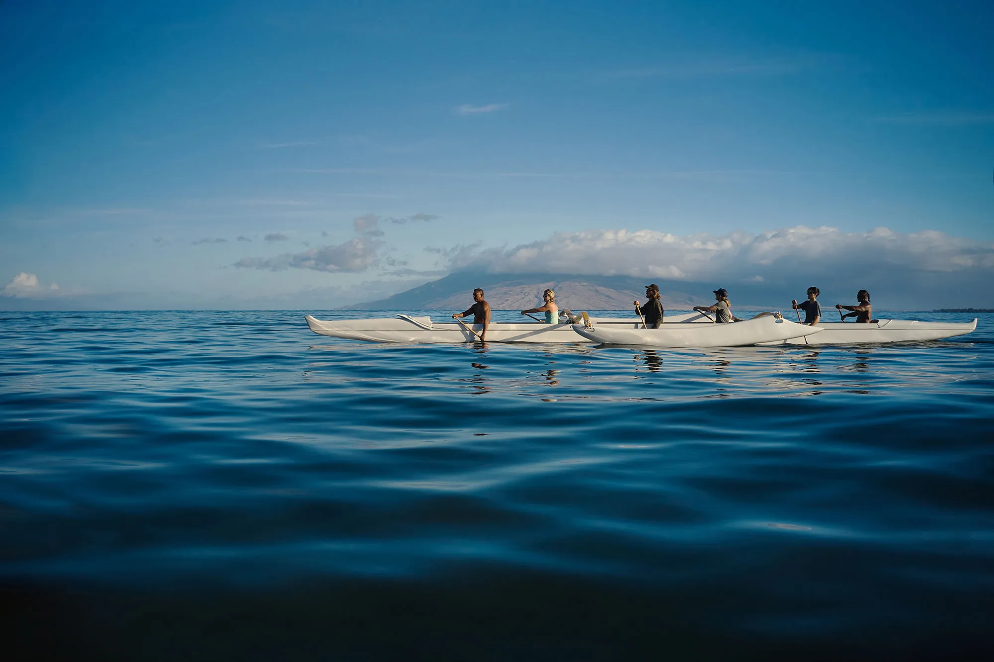A group of people canoeing in traditional Hawaiian canoes over a calm ocean with misty mountains in the background