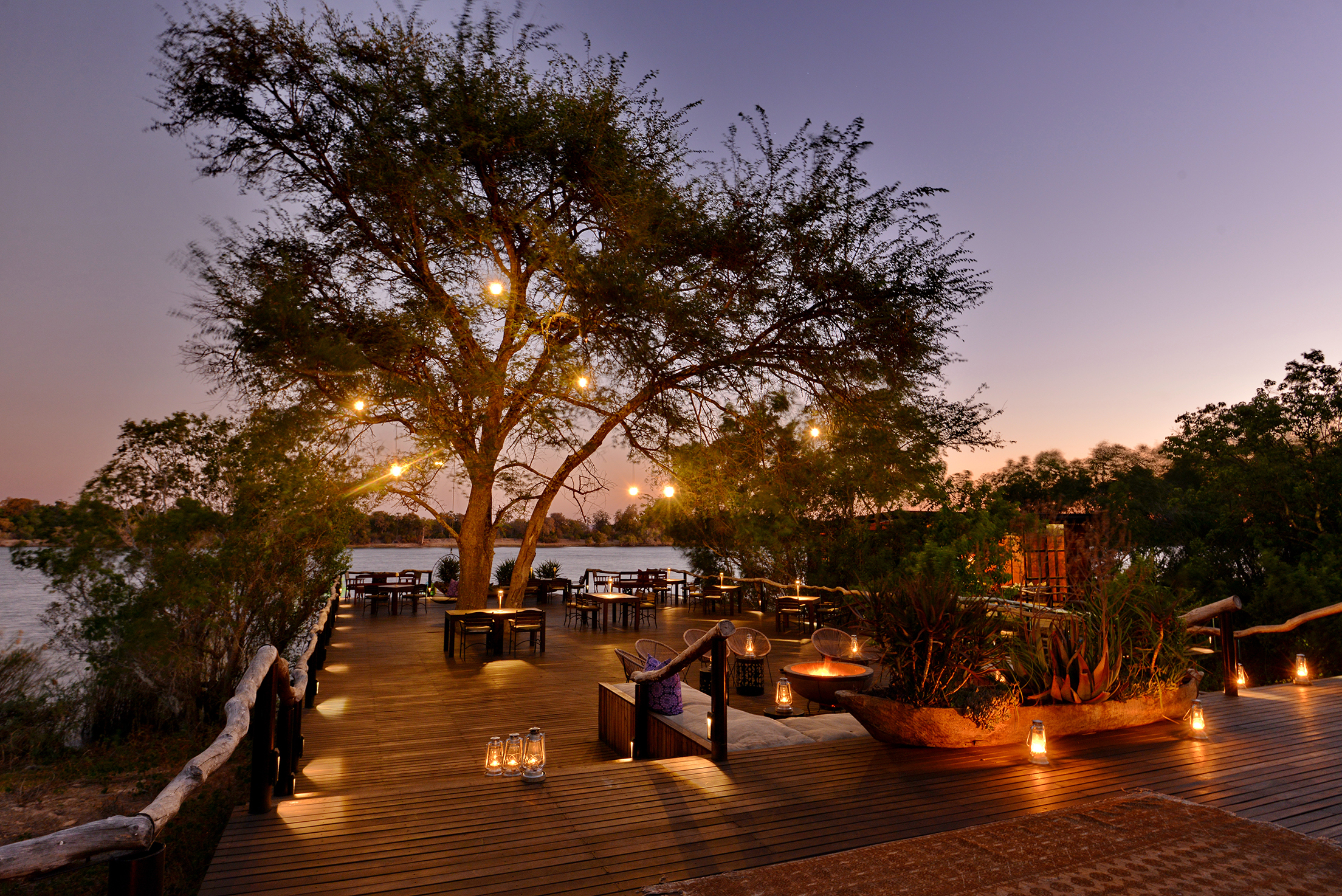 Serene outdoor dining and lounge area at Mukwa River Lodge with wooden decking, ambient tree lighting, and a river view at sunset.