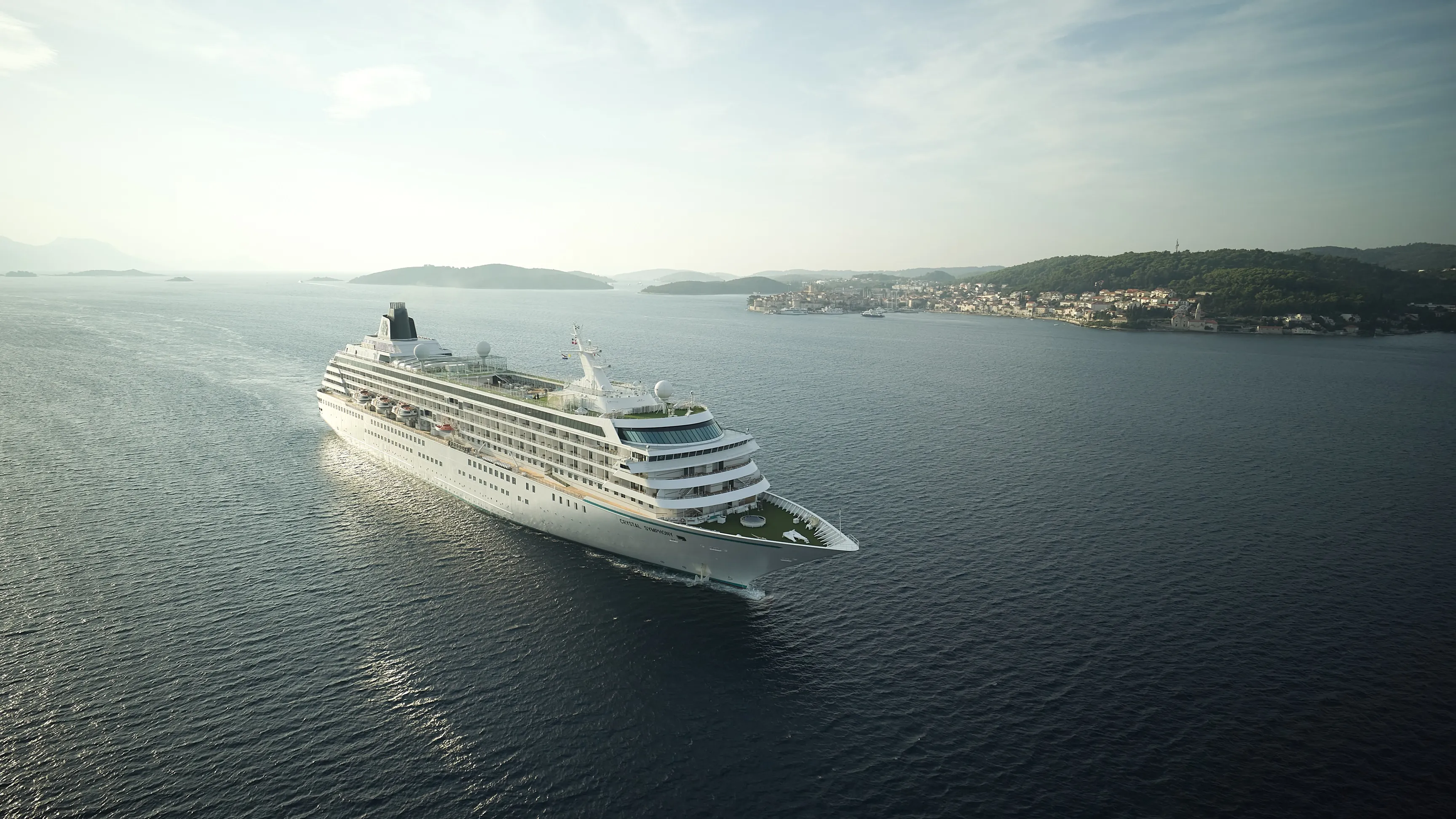 Crystal Symphony cruise ship sailing through calm waters with a scenic coastline and hills in the distance.