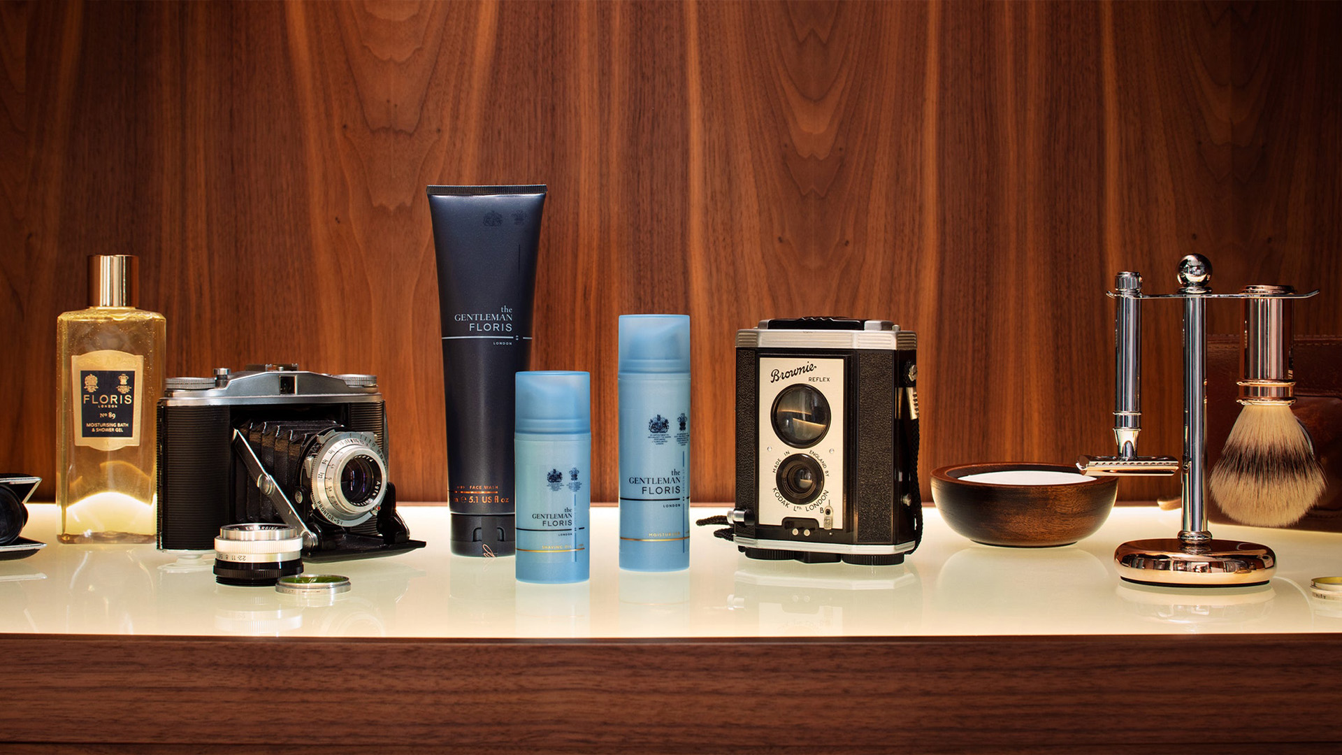 A men's grooming kit of shaving cream and balm, razor and brush and aftershave is staged next two 60s style cameras
