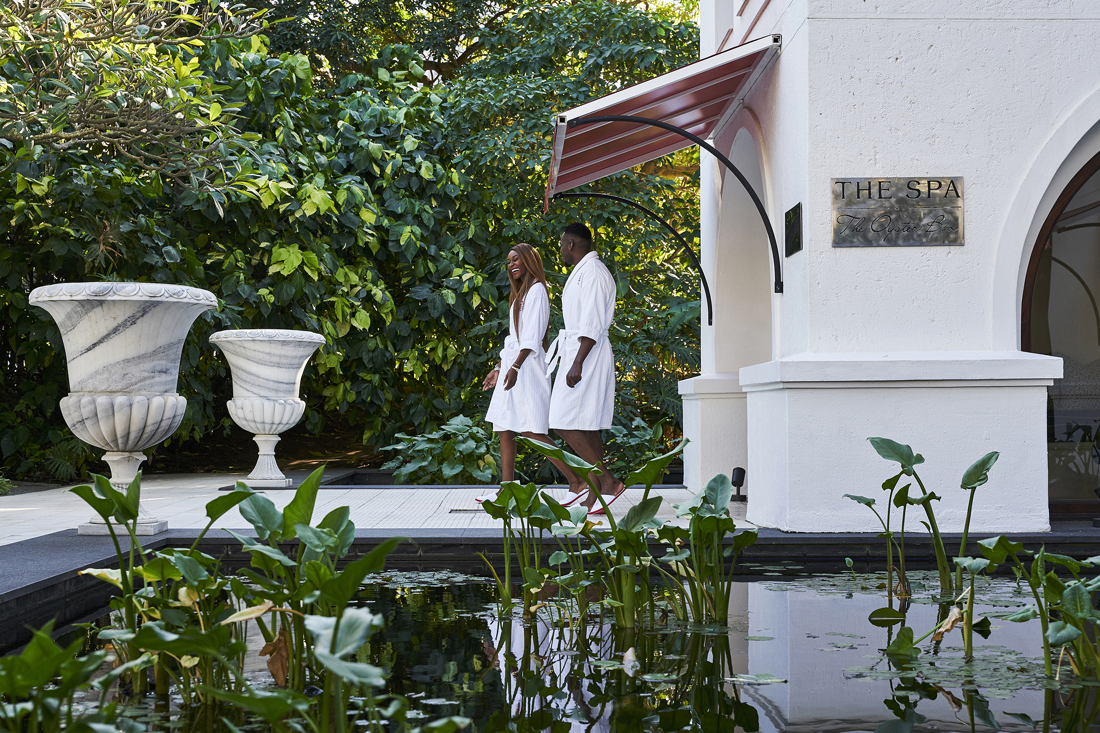 Two people in white bath robes walking out of the spa beside a pond with large vases