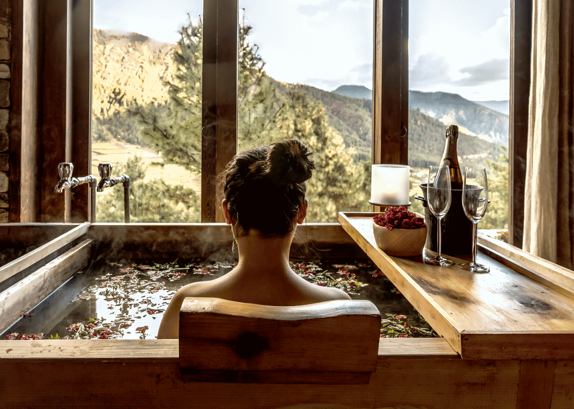Person soaking in a wooden hot bath with floating botanicals, overlooking a scenic mountain landscape through large windows.