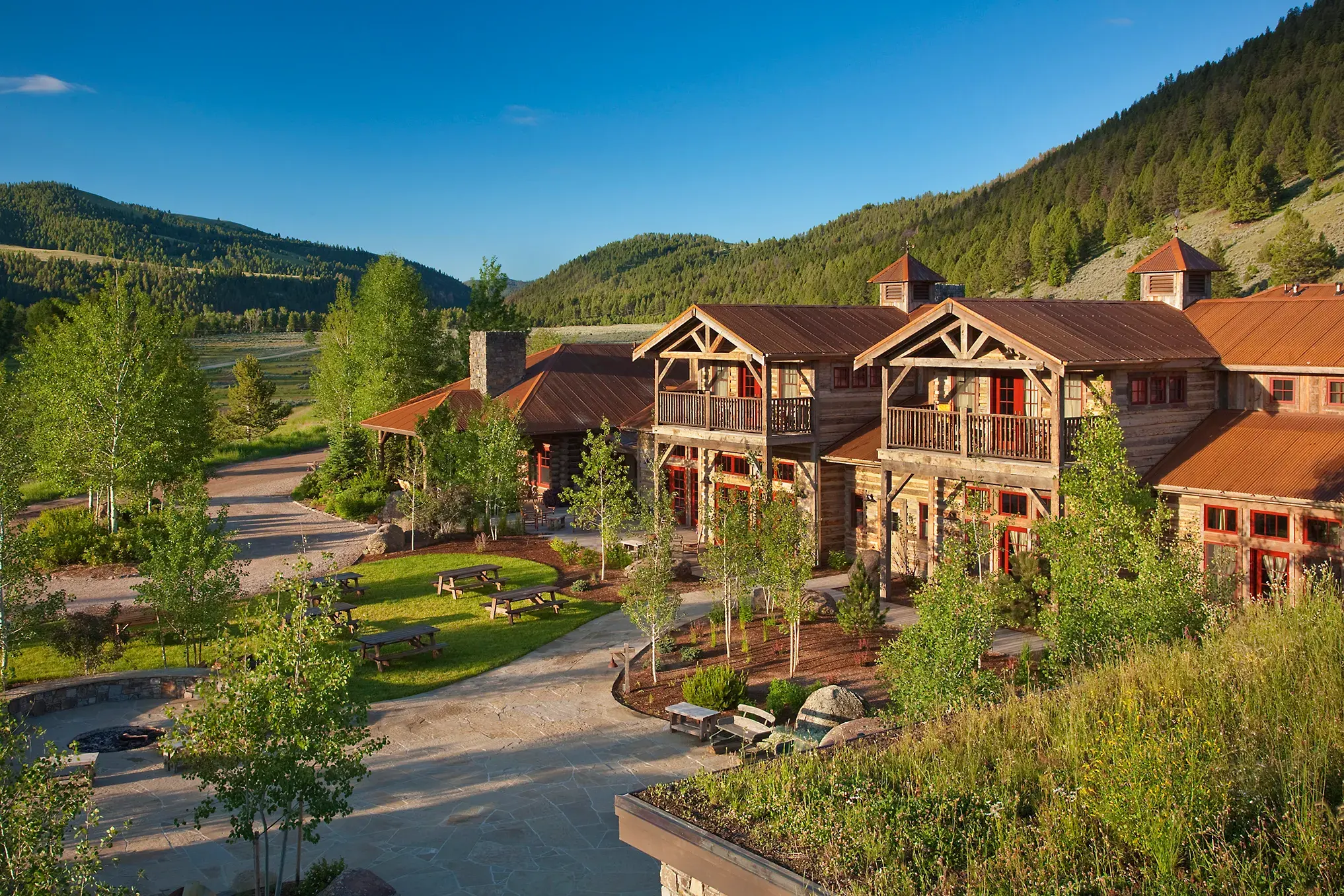 The large wooden exterior of the Granite Lodge at The Ranch at Rock Creek in Montana with balconies sitting among trees with green mountains behind under a clear sky