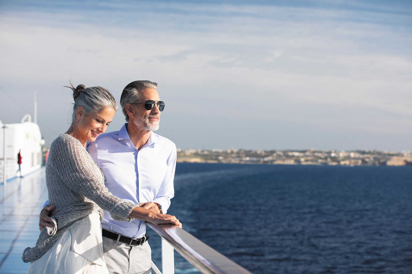 A middle-aged man and woman stand on a cruise ship deck overlooking the sea, smiling as they hold hands.