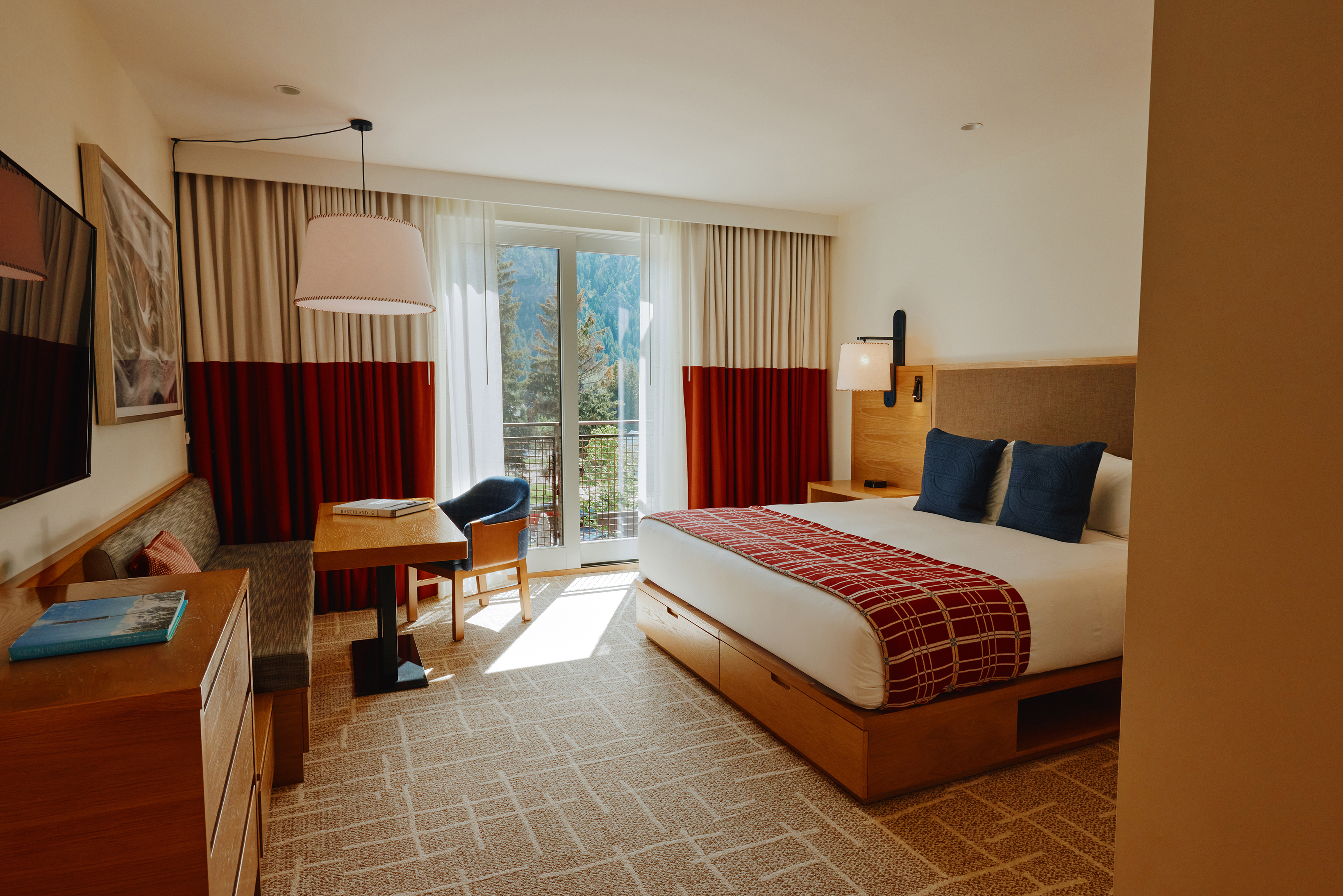 A modern hotel room at Limelight Ketchum featuring a large bed with red and white bedding, blue pillows, a desk and chair, wall-mounted TV and floor-to-ceiling windows with matching curtains
