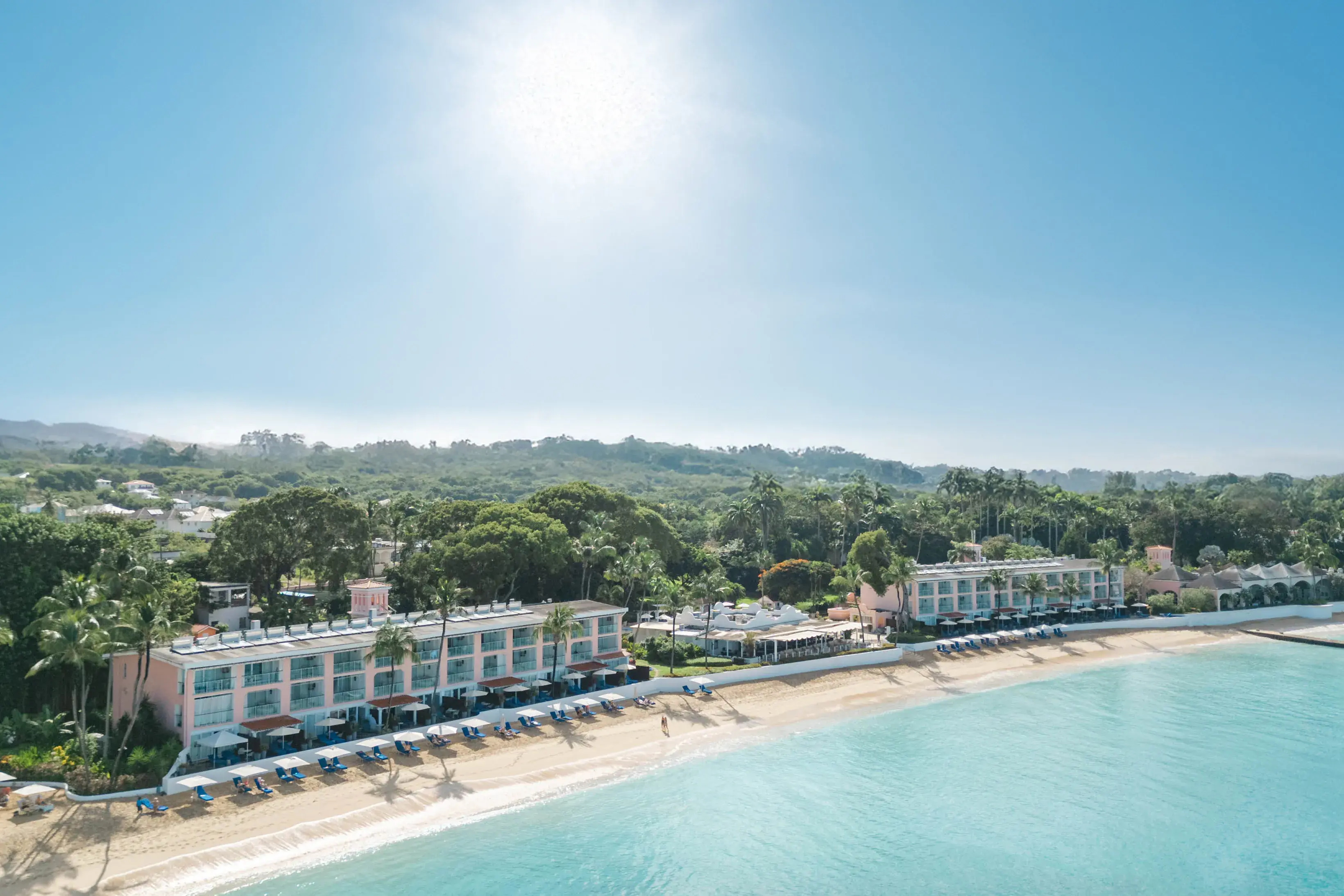 An aerial view of Fairmont Royal Pavilion in Barbados from the beach featuring hotel buildings lining the sand in front of tropical trees
