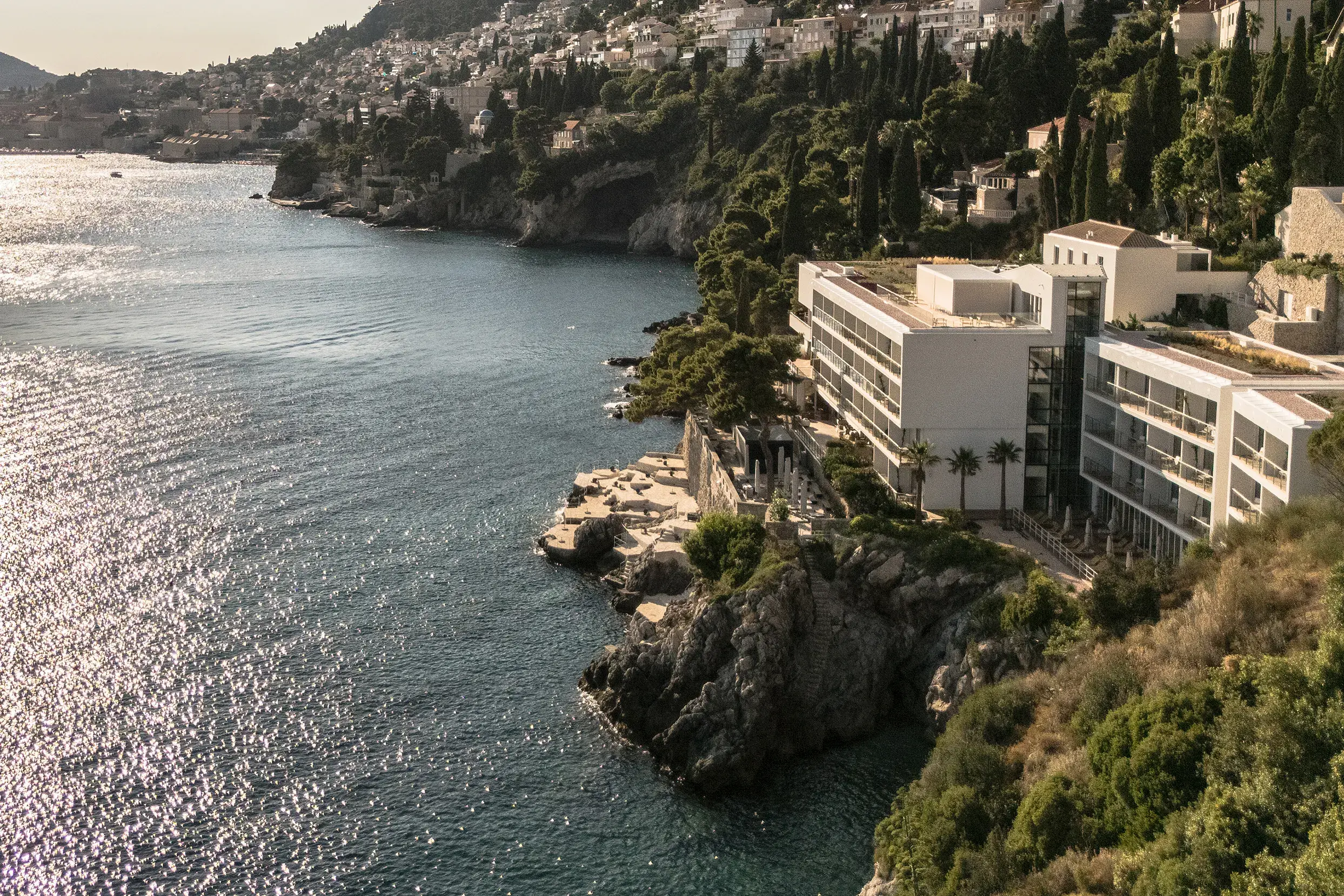 Exterior aerial view of Villa Dubrovnik featuring the building surrounded by trees and the beach area by the water, with more building and the Old Town behind