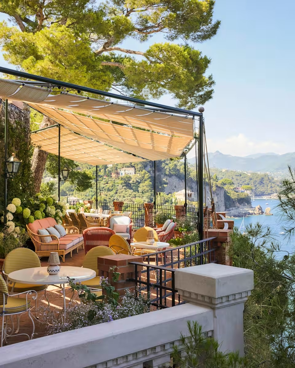 Elegant outdoor terrace at Belmond Villa Bertrice with colourful seating, shaded canopy, and panoramic view of lush hills and blue Mediterranean coastline.
