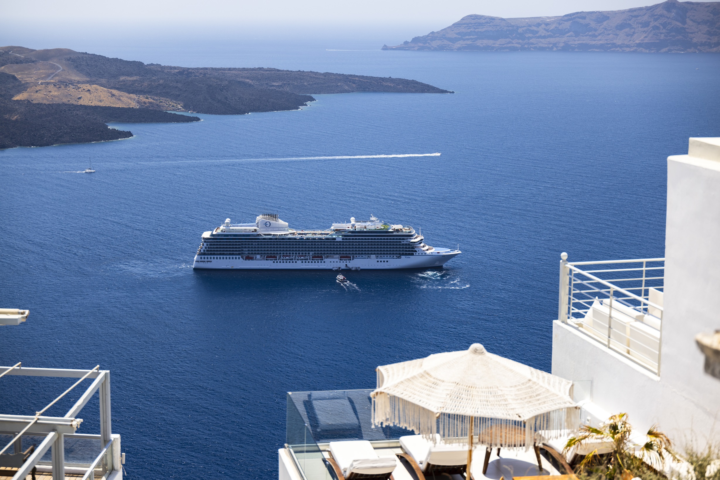 Oceania Cruises ship sailing near a mountainous Santorini coast, viewed from a balcony with white railings and an umbrella.