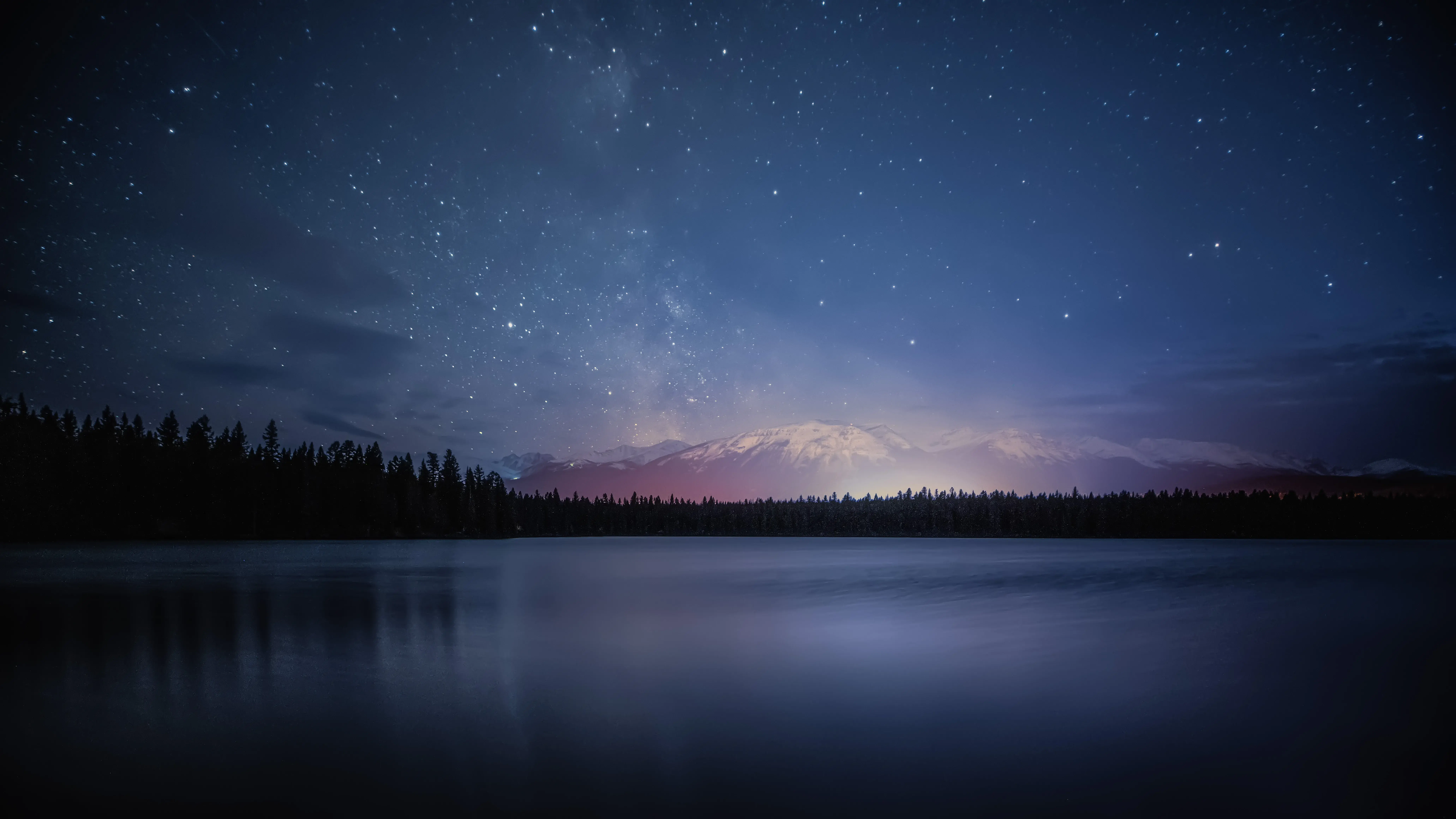 Starry night sky over snow-capped mountains in Jasper National Park reflecting on a calm lake with a silhouette of pine trees.