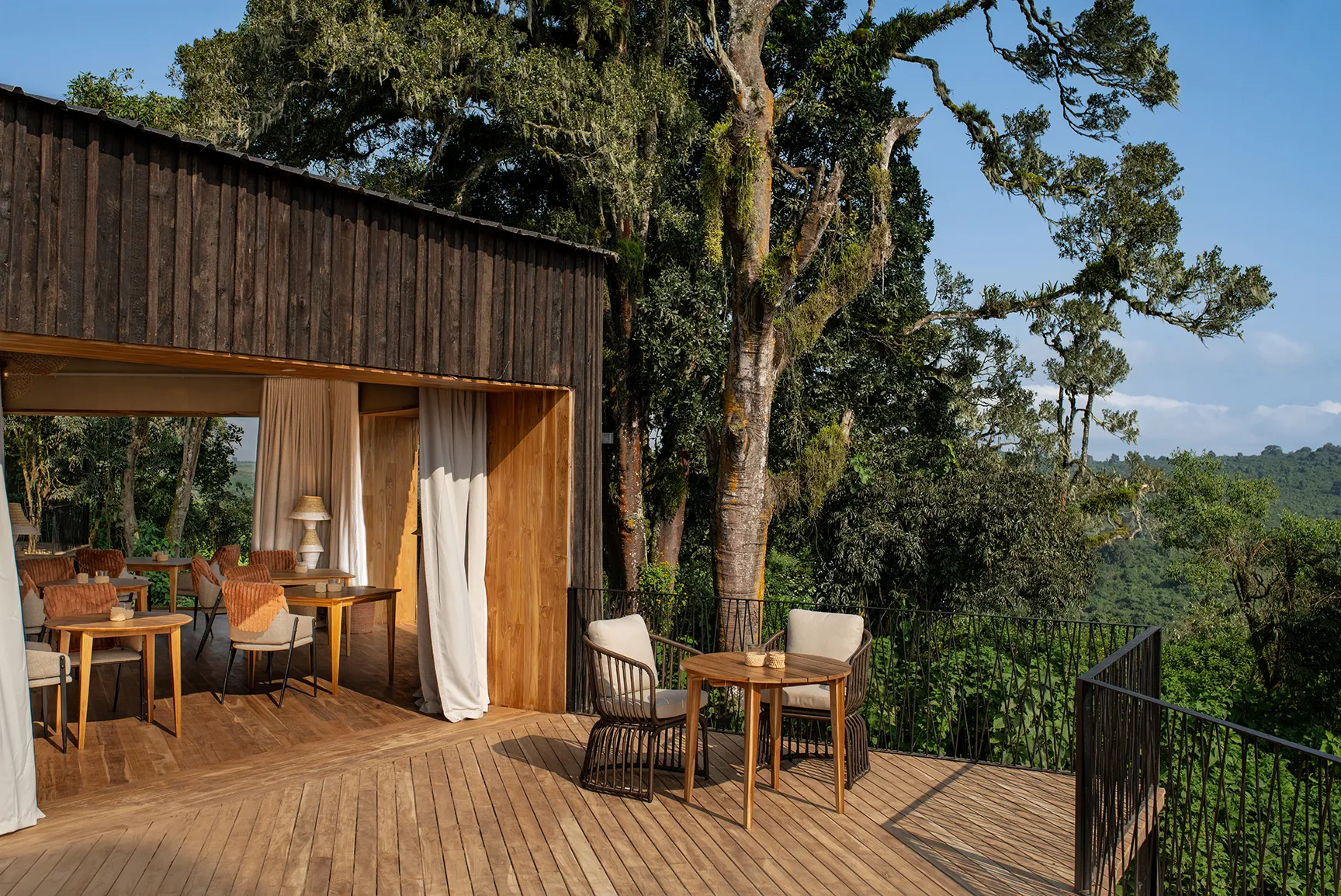 Open-air lodge dining terrace with wooden tables and chairs overlooking a lush forest landscape