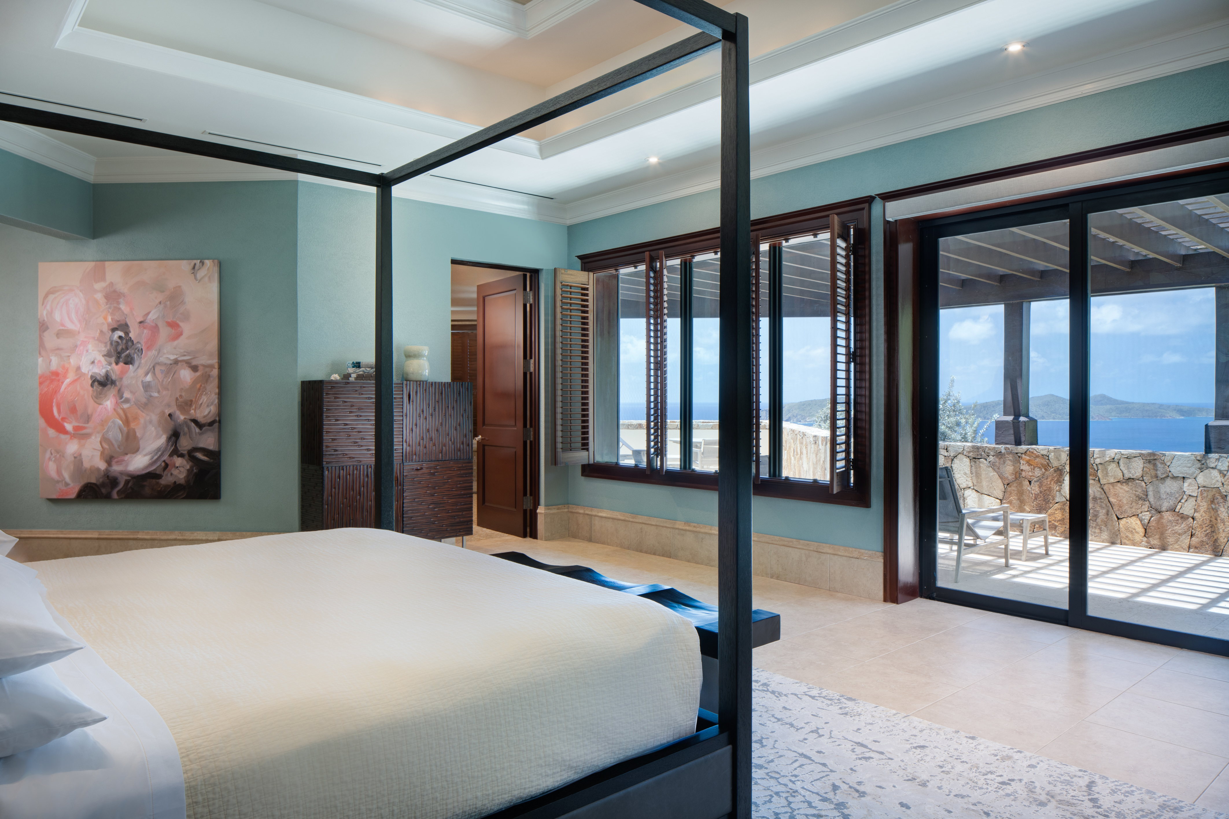 Four poster bed with white sheets in a blue-walled room with floral artwork and sliding glass doors to a terrace