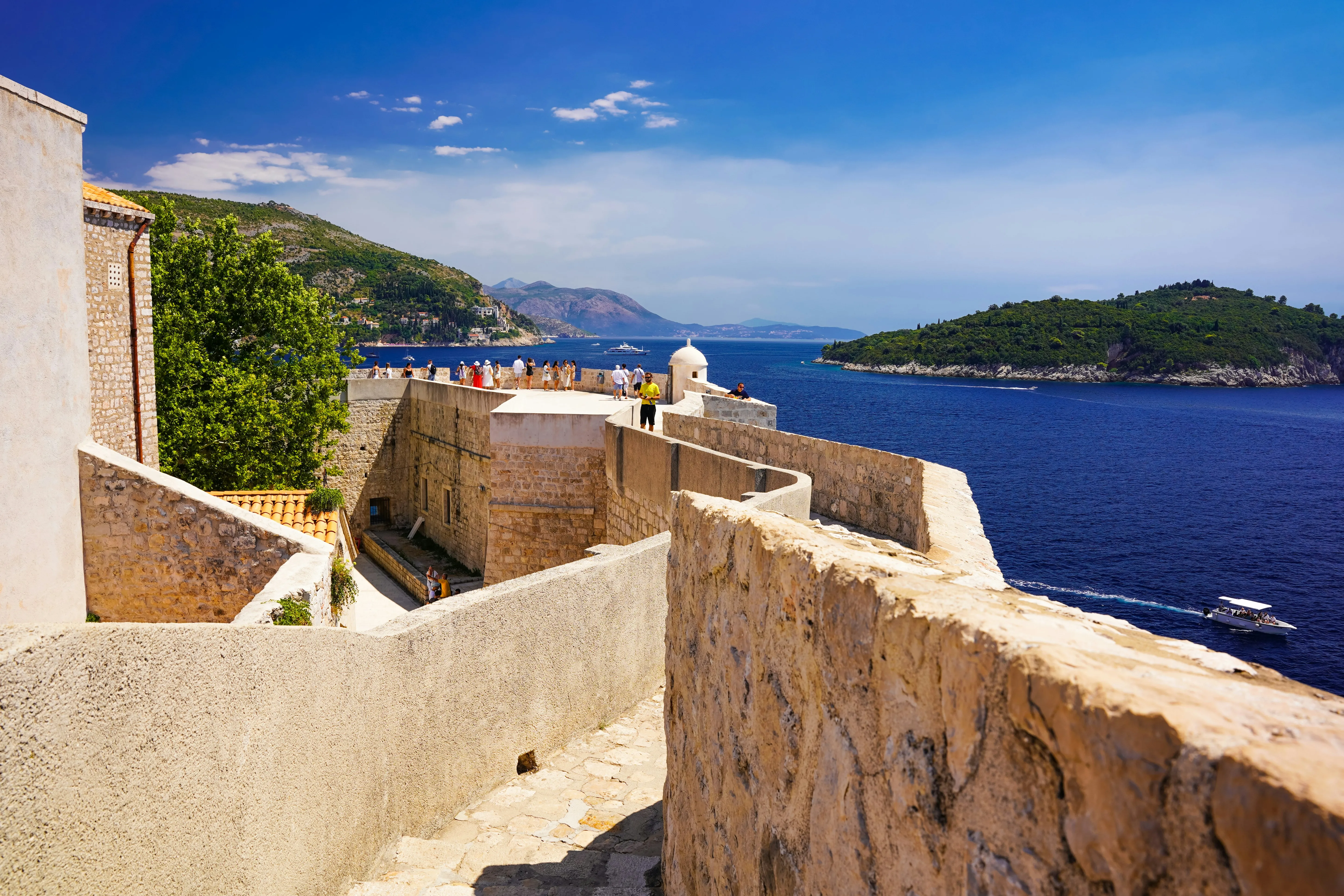 View down the pathway along Dubrovnik's stone city walls with buildings on one side and blue ocean, a boat and islands on the other beneath a clear sky