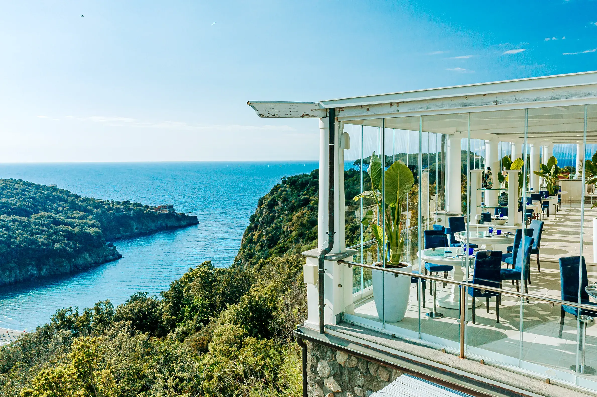 San Montano Resort on Ischia with a stunning view of the Mediterranean Sea and lush green hills, featuring an elegant dining area with glass walls and blue chairs.