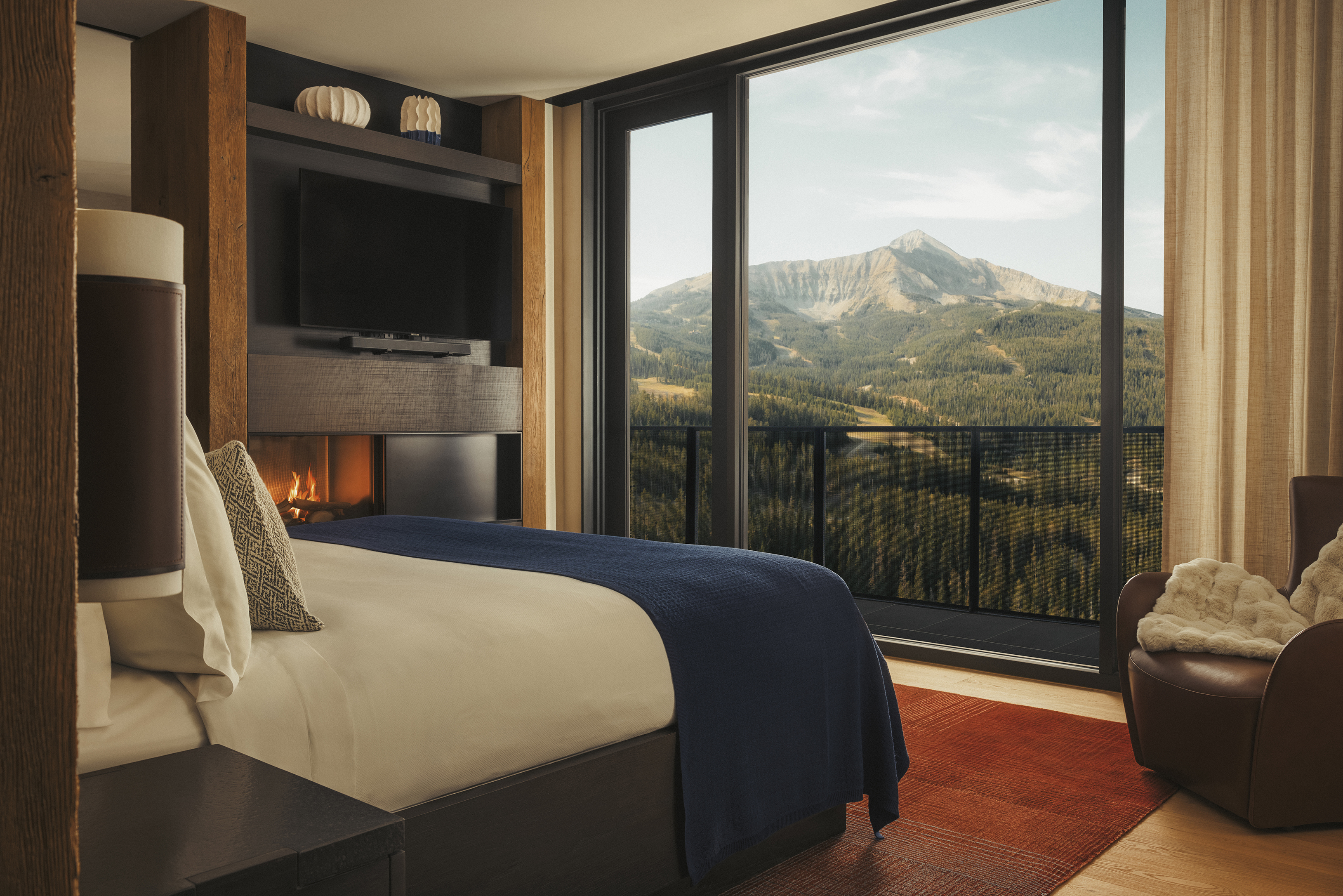 A cosy and modern bedroom featuring a lit fireplace and bed with blue covers facing large windows looking out to a mountain beyond