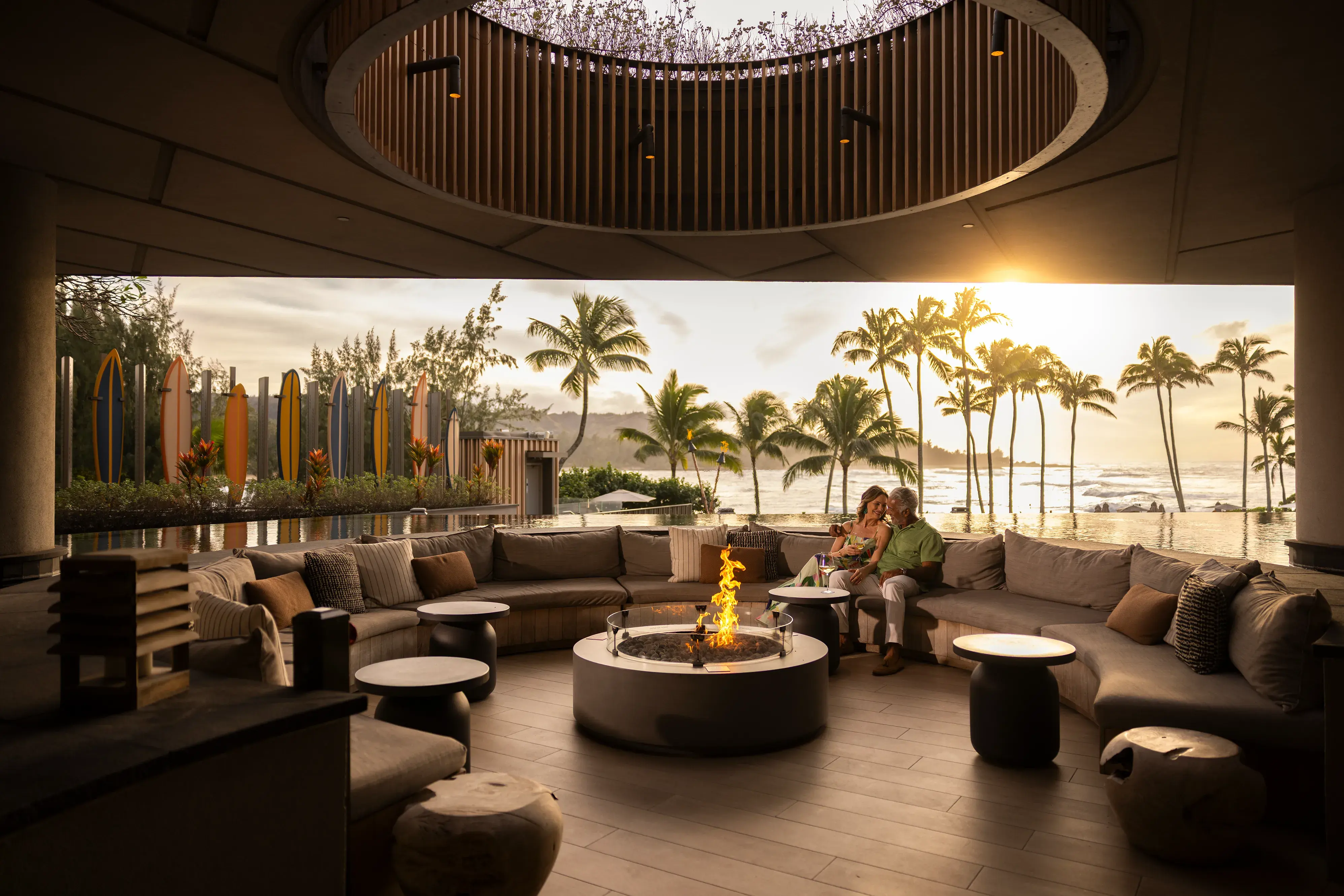 Two people lounging in the Oculus At Off The Lip bar at Ritz-Carlton Turtle Bay with sofas circling a firepit backed by views out to sea with palms and surfboards 