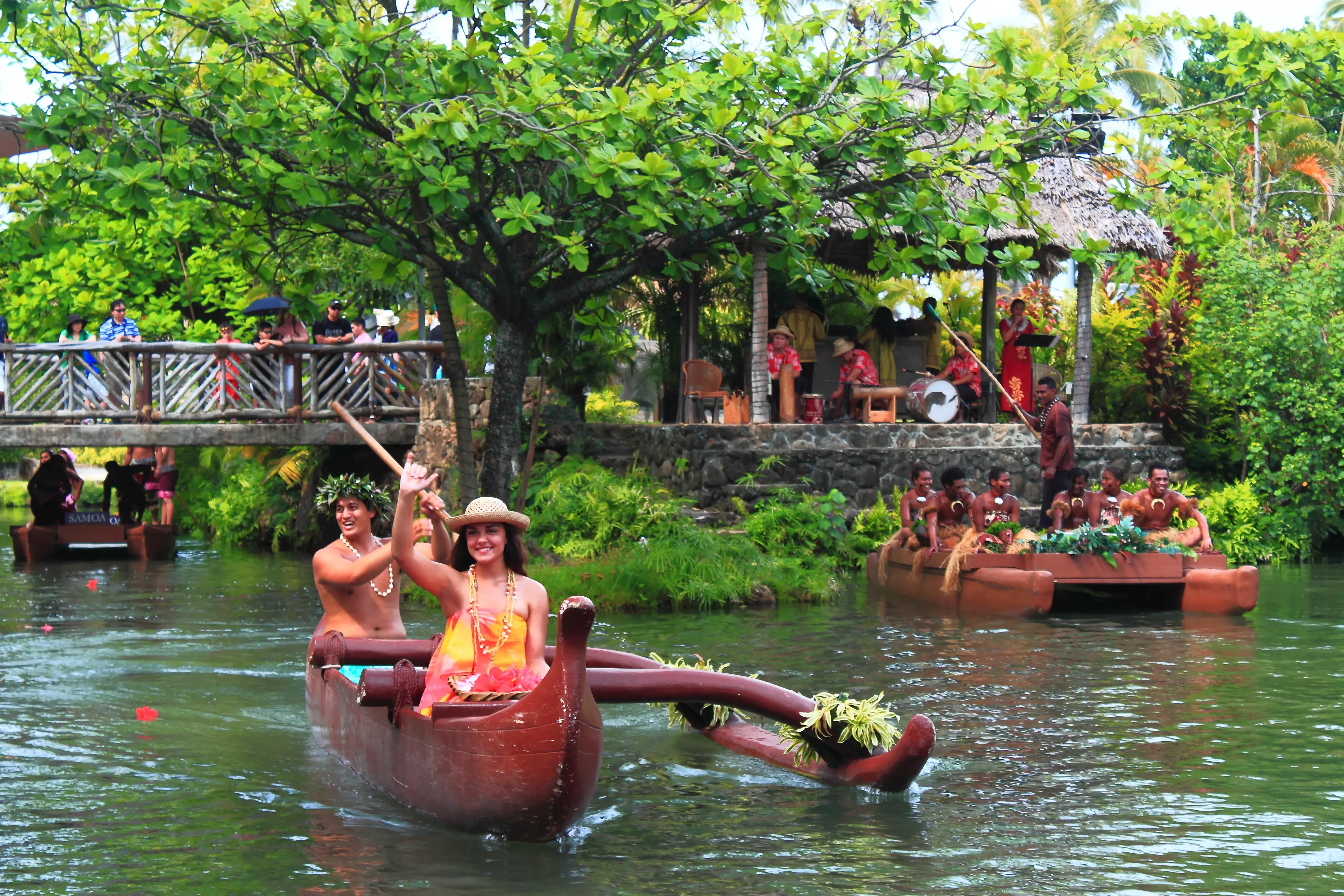 People in a traditional Polynesian canoe with a live band on land behind at the Polynesian Cultural Center