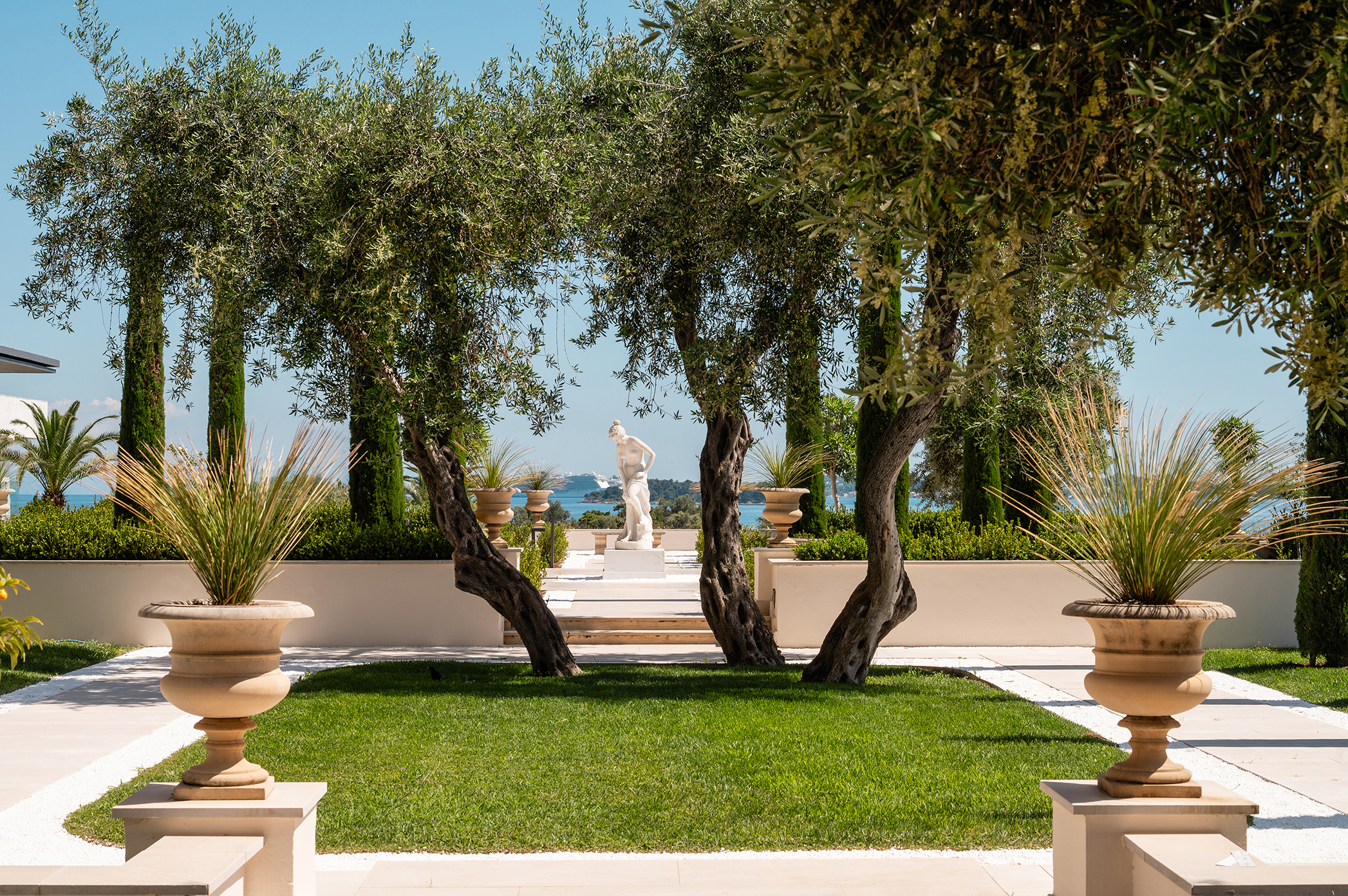 Elegant garden view at Grecotel Corfu Imperial featuring lush greenery, classical statues, and a serene sea backdrop.