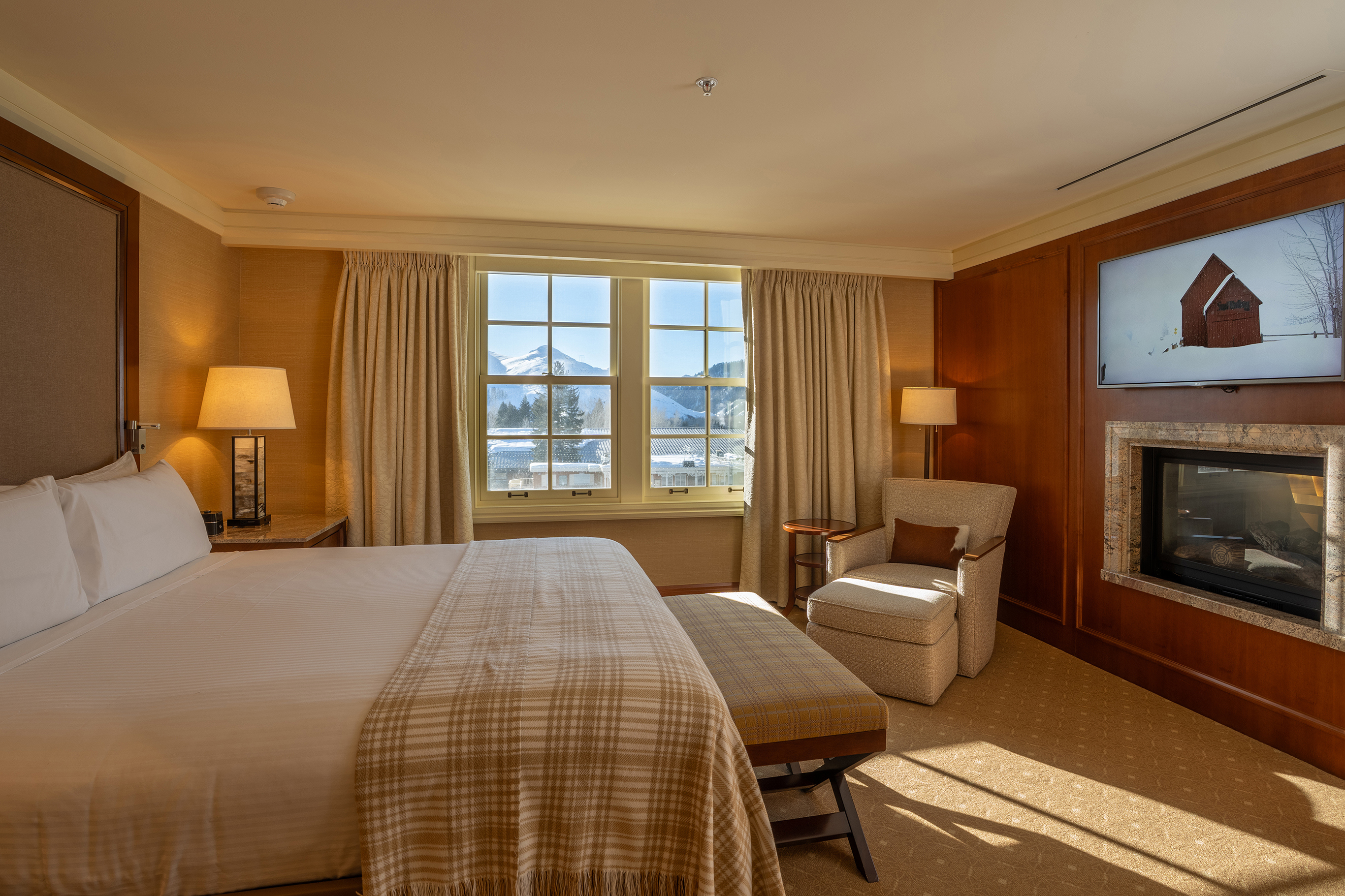 A cosy hotel suite at Sun Valley Lodge featuring a large bed, armchair with ottoman, fireplace, and a window with mountain views