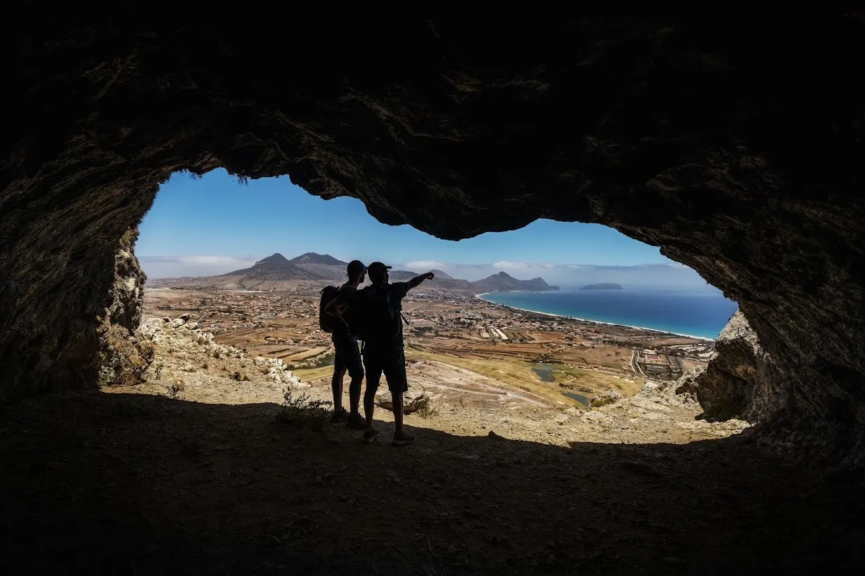 Silhouetted hikers standing inside a cave, overlooking a coastal landscape with mountains and the ocean.